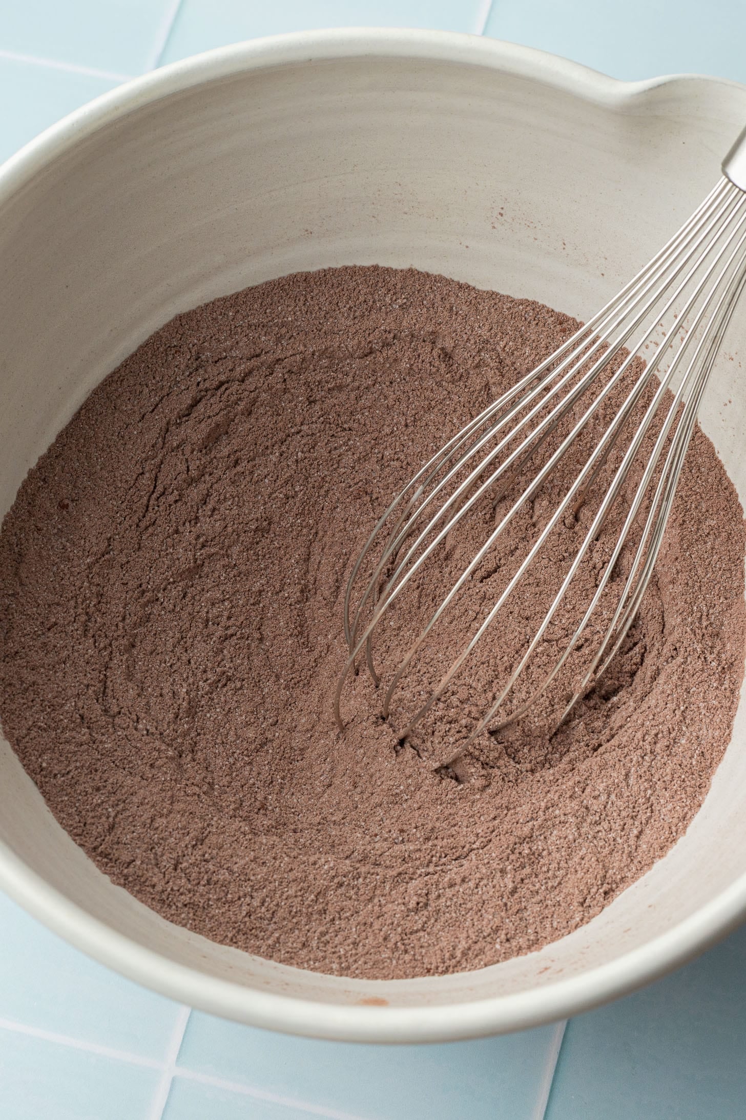 A white bowl filled with cocoa powder sits on a light blue countertop. A metal whisk is partially submerged in the powder, ready for mixing.