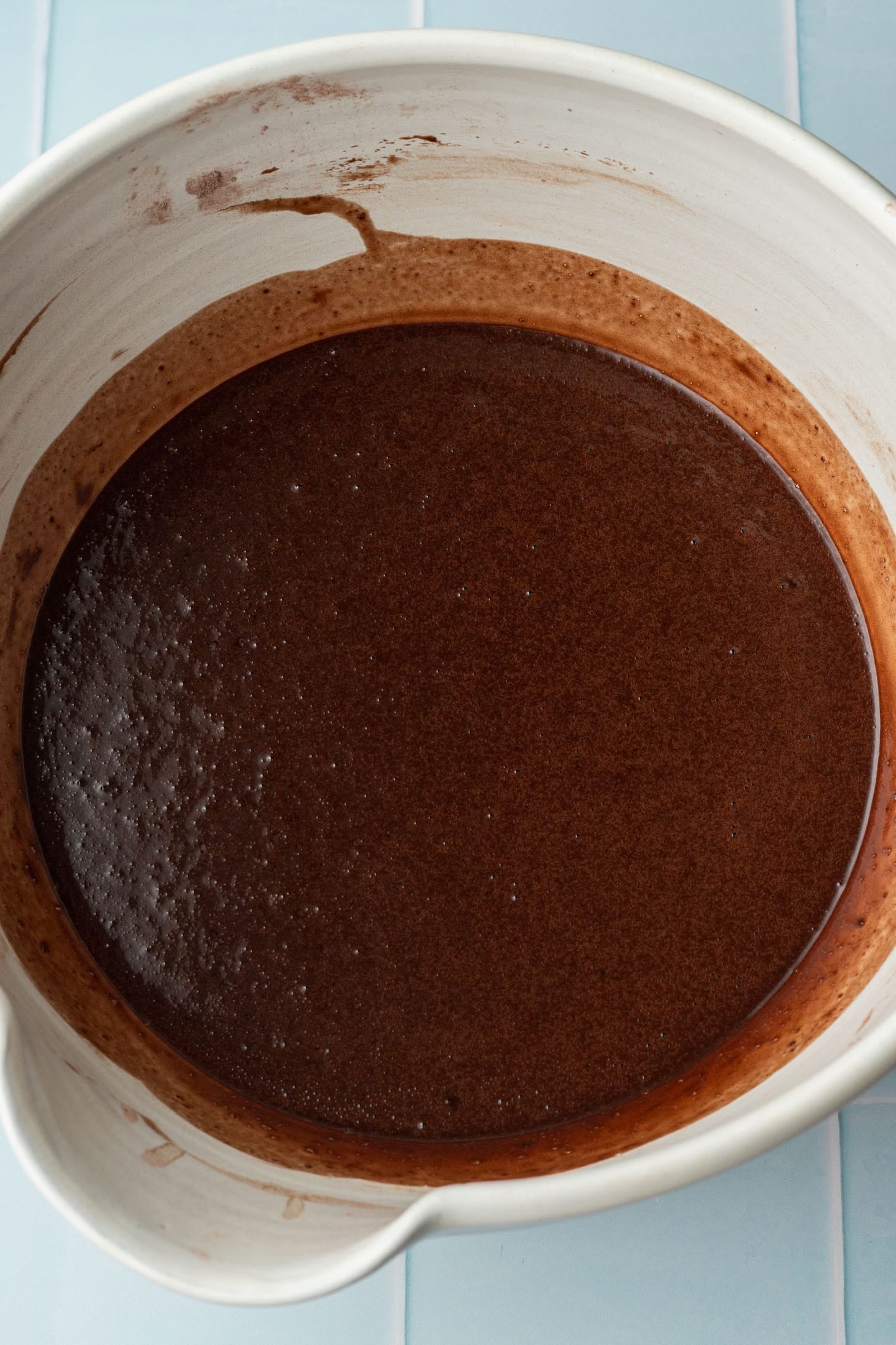 A white mixing bowl filled with smooth, dark chocolate cake batter on a light blue countertop, suggesting home baking in progress.