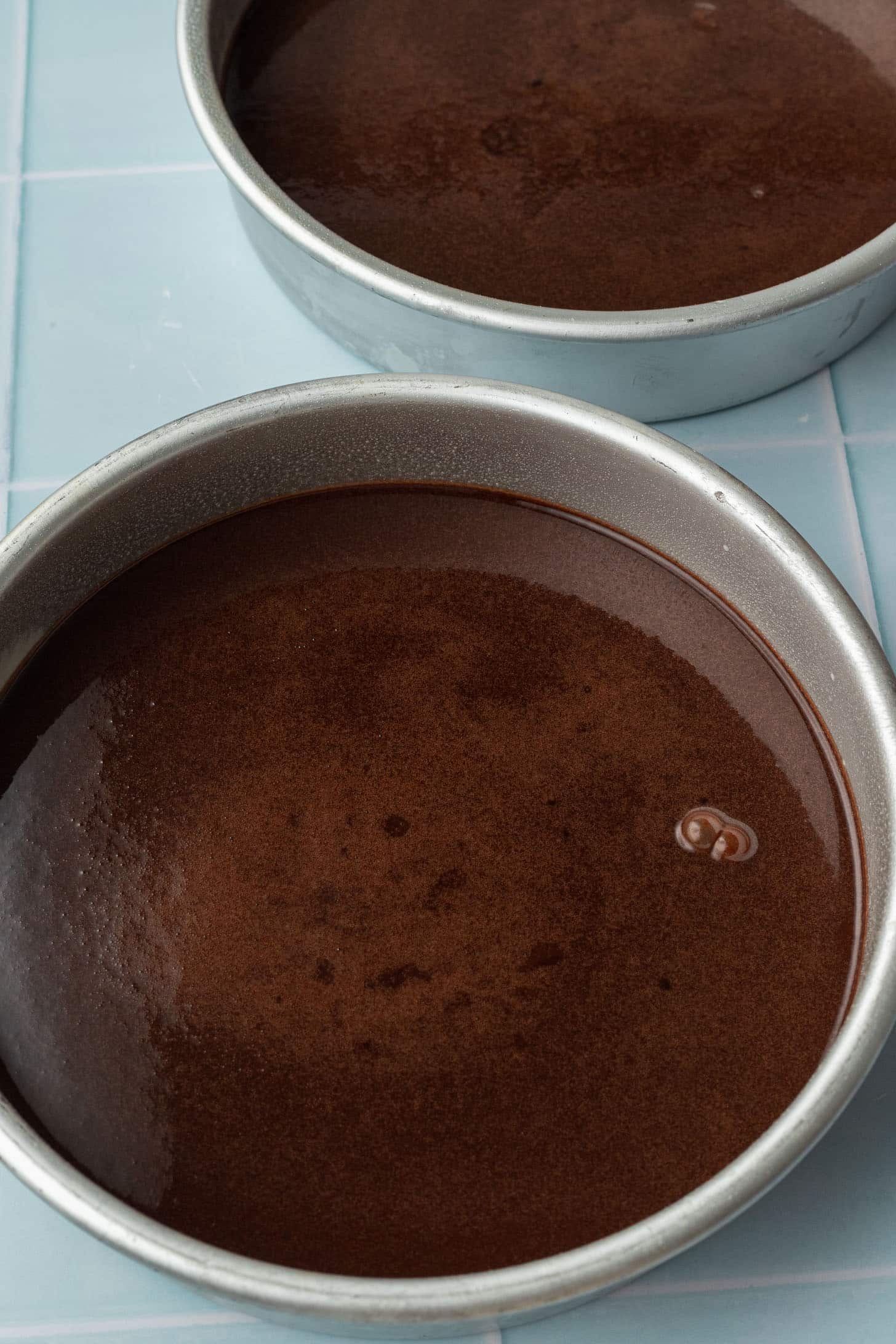 Two round cake pans filled with smooth, dark chocolate batter are placed on a light blue tiled surface, ready for baking.