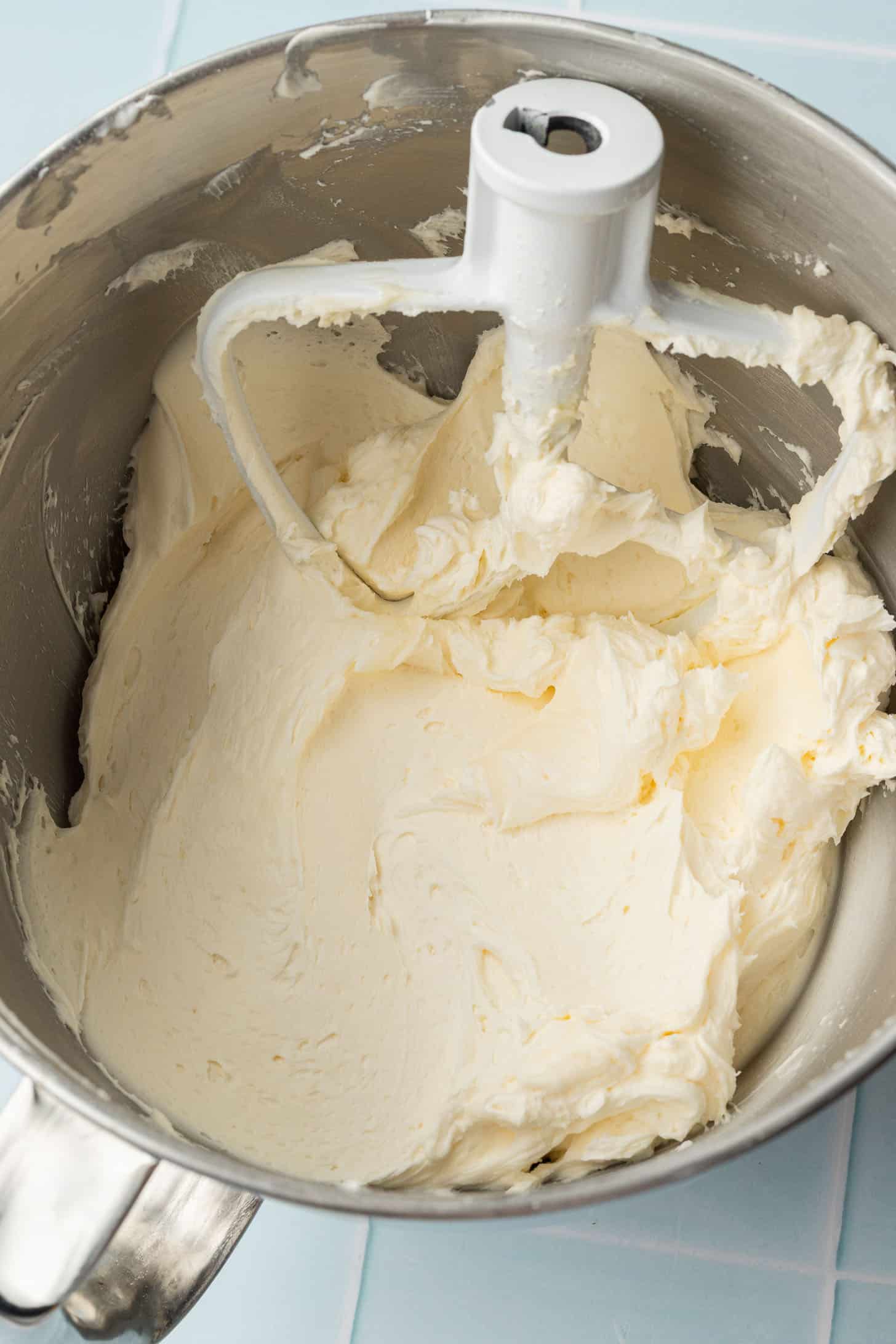 Creamy, pale yellow batter is mixed in a stainless steel bowl with a paddle attachment, sitting on a light blue tiled surface.