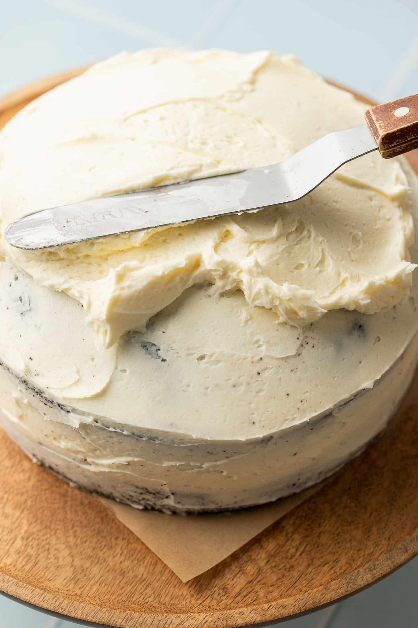 A cake being frosted with creamy white icing using an offset spatula on a wooden board. The scene conveys a sense of home baking and preparation.
