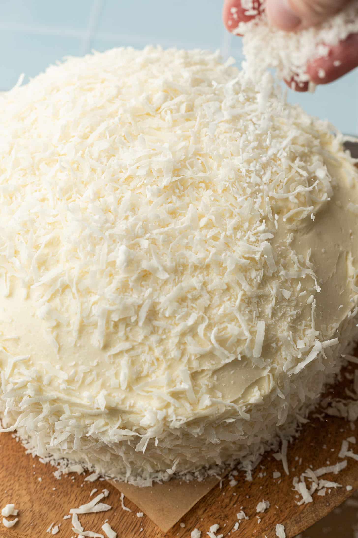 A hand sprinkles shredded coconut over a round cake with white icing, creating a textured, snowy appearance. The scene conveys freshness and indulgence.