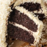 A rich chocolate cake with creamy white frosting and coconut shavings, partially sliced, displayed on a wooden board. The texture looks moist and indulgent.