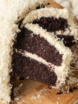 A rich chocolate cake with creamy white frosting and coconut shavings, partially sliced, displayed on a wooden board. The texture looks moist and indulgent.