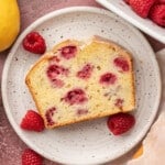 Slice of raspberry lemon loaf on a speckled plate, surrounded by fresh raspberries and whole lemons. Nearby, a dish holds more raspberries.