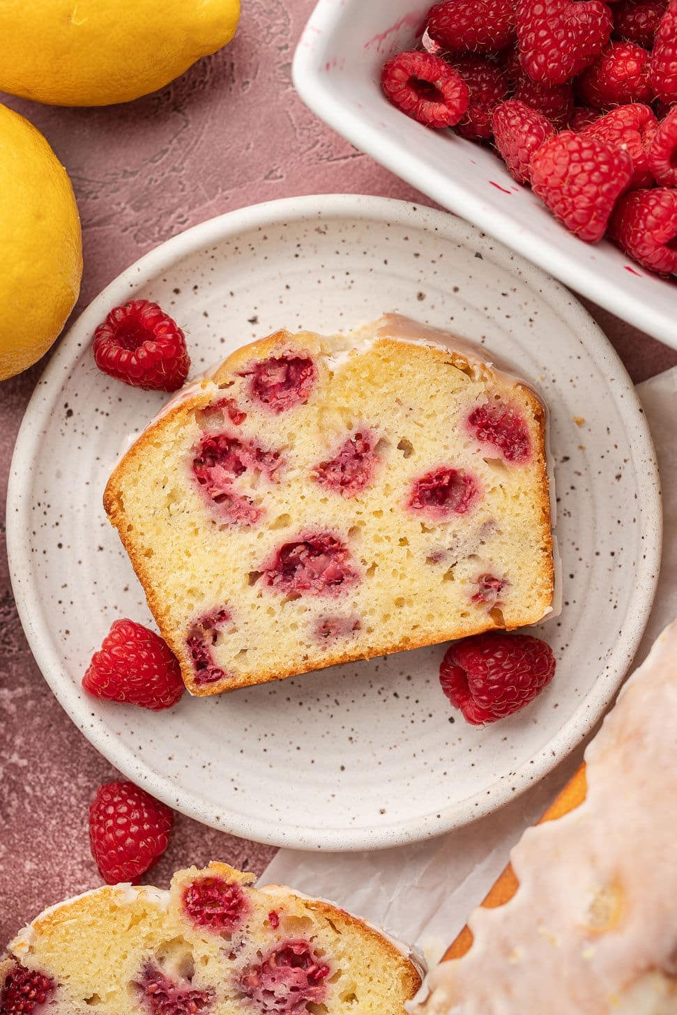 Slice of raspberry lemon loaf on a speckled plate, surrounded by fresh raspberries and whole lemons. Nearby, a dish holds more raspberries.