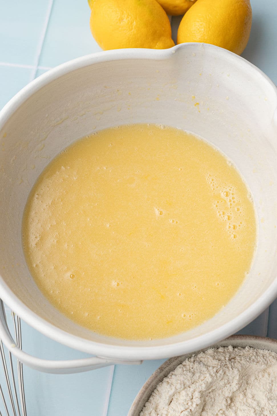 A bowl of lemon batter sits on a light blue tiled surface, with three whole lemons in the background and a bowl of flour nearby, ready for baking.