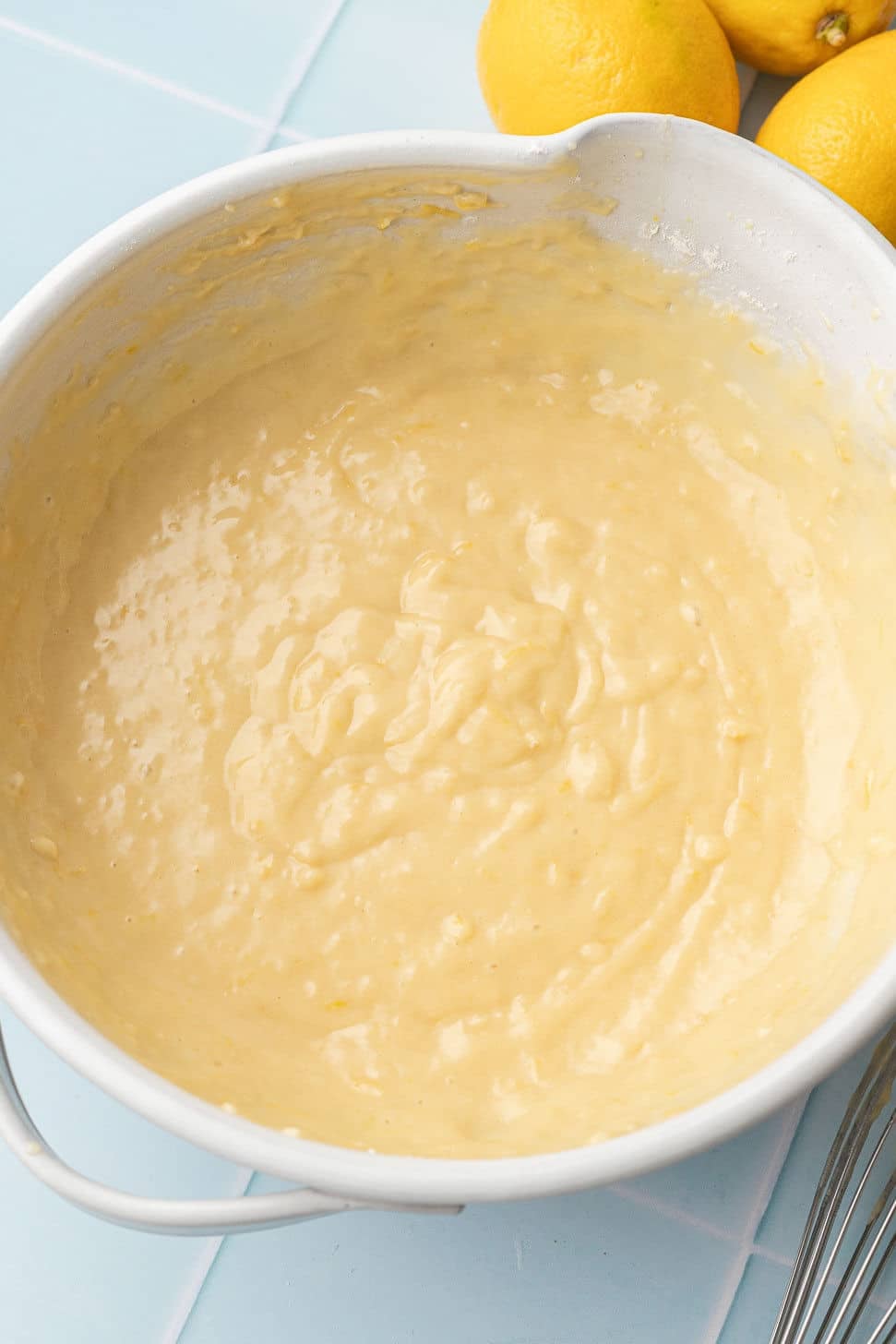 A mixing bowl filled with smooth, creamy lemon batter sits on a light blue countertop. Bright yellow lemons lie adjacent, suggesting a fresh, zesty flavor.