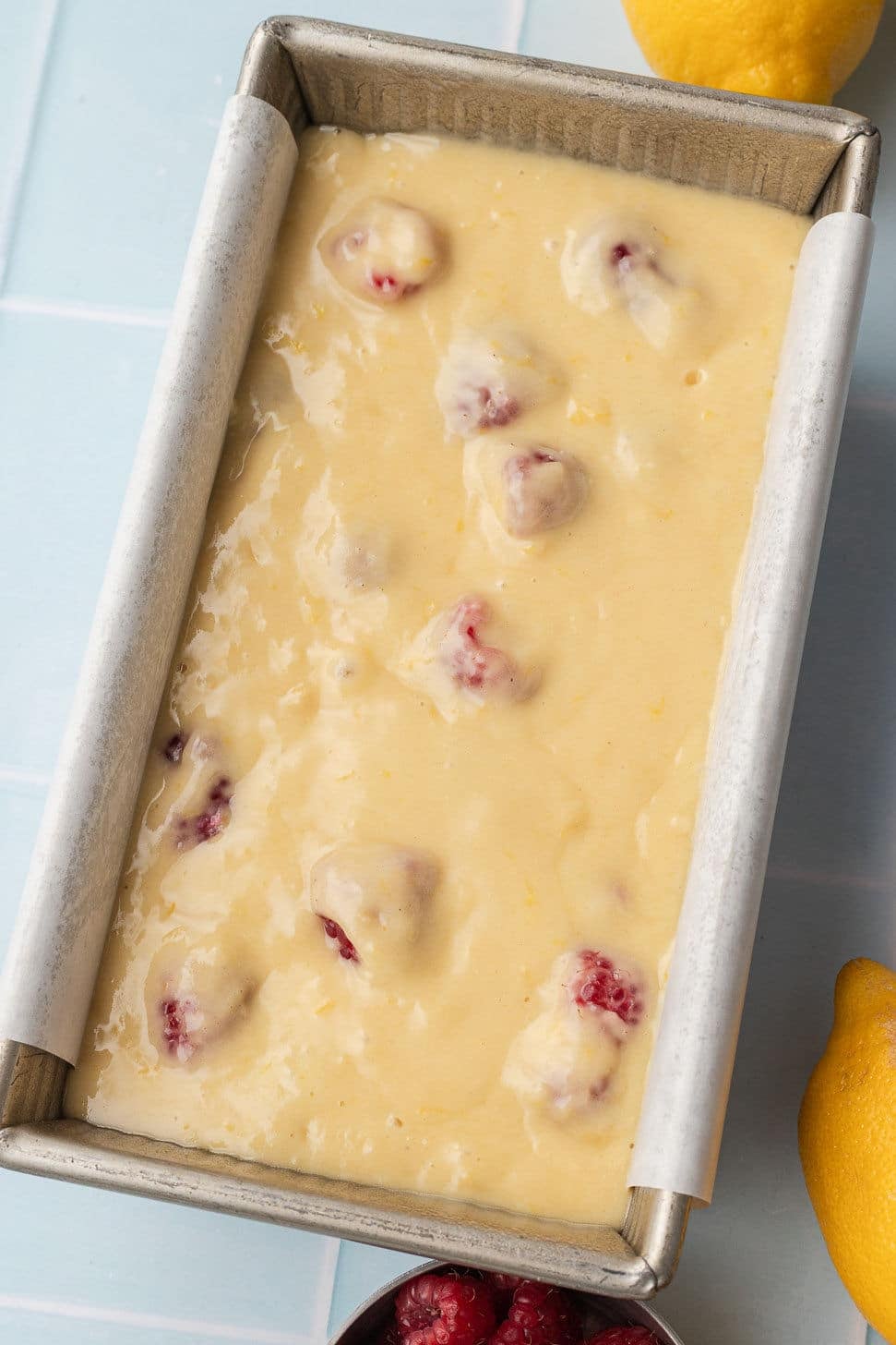 Loaf pan with creamy batter mixed with raspberries, on a light blue surface. Lemons and raspberries surround the pan, creating a bright, fresh feel.