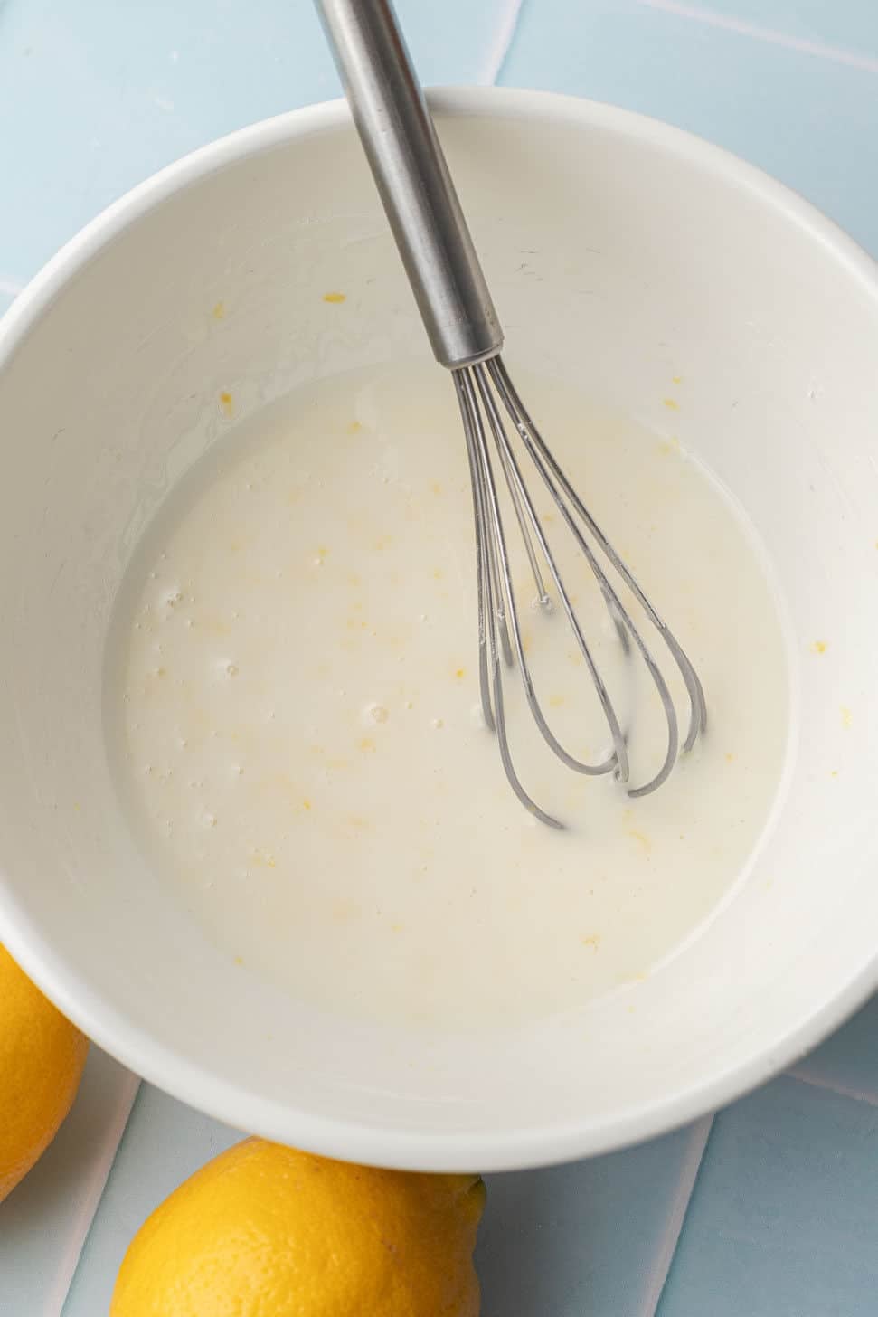 A white bowl with a metal whisk is mixing a lemon-flavored batter. Two whole lemons are placed nearby on a pastel blue surface.