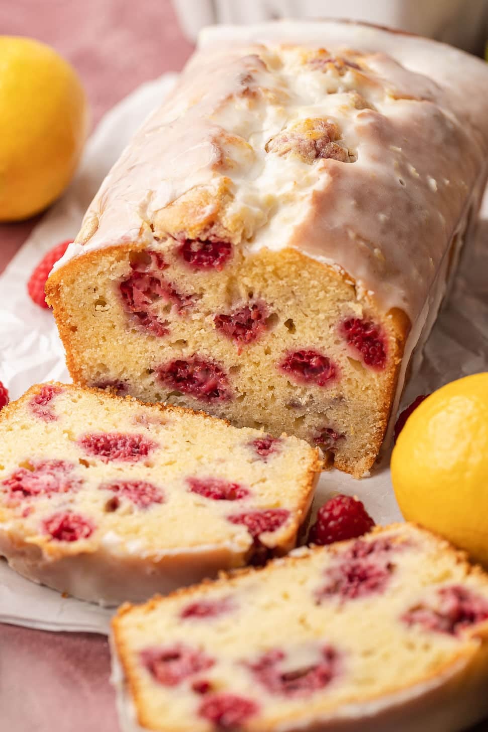 Lemon raspberry loaf cake with glaze, sliced to show moist crumb and vibrant raspberries, surrounded by fresh lemons and a blurred background.