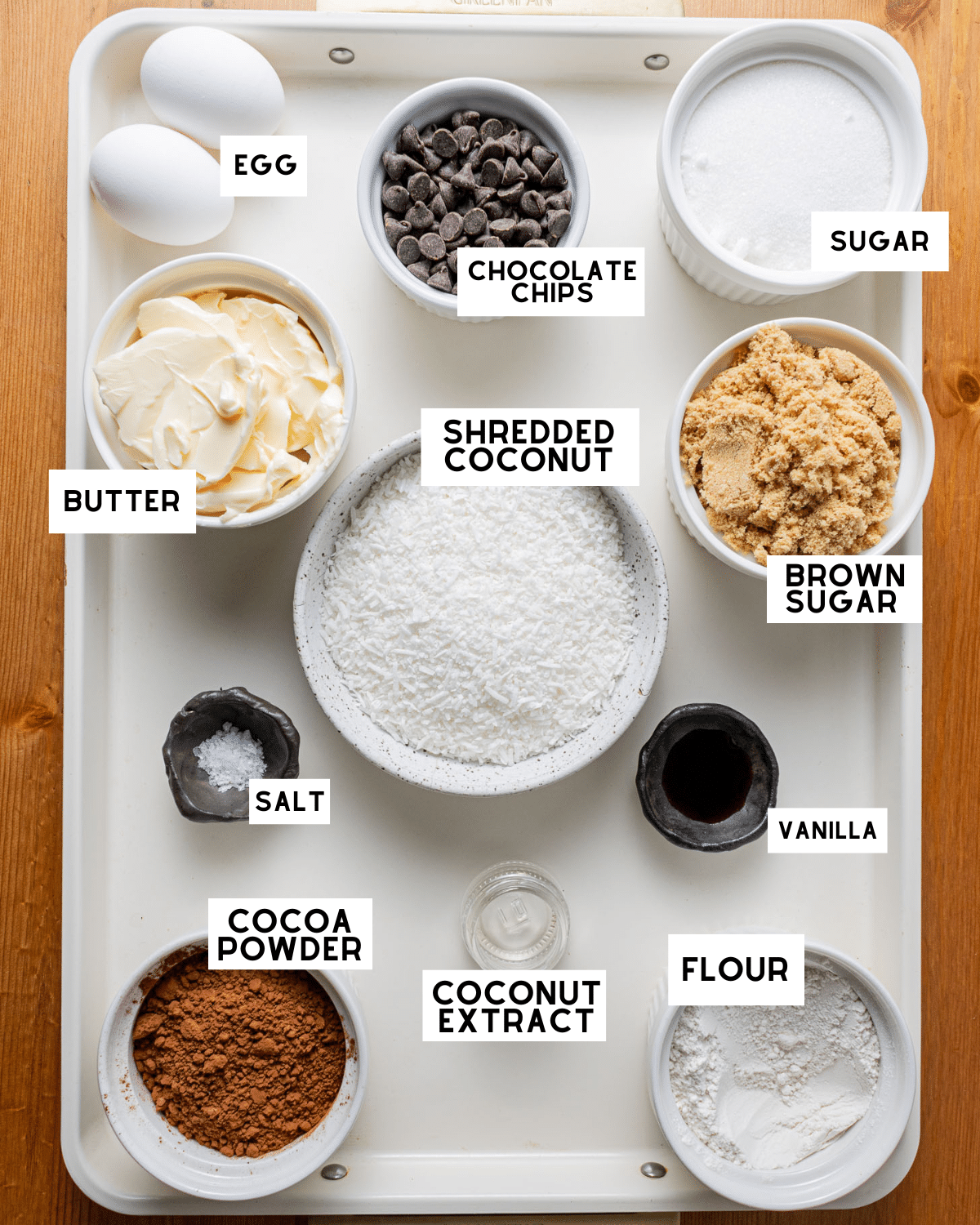 Tray with baking ingredients: eggs, butter, shredded coconut, chocolate chips, cocoa powder, flour, sugar, brown sugar, salt, vanilla, coconut extract.