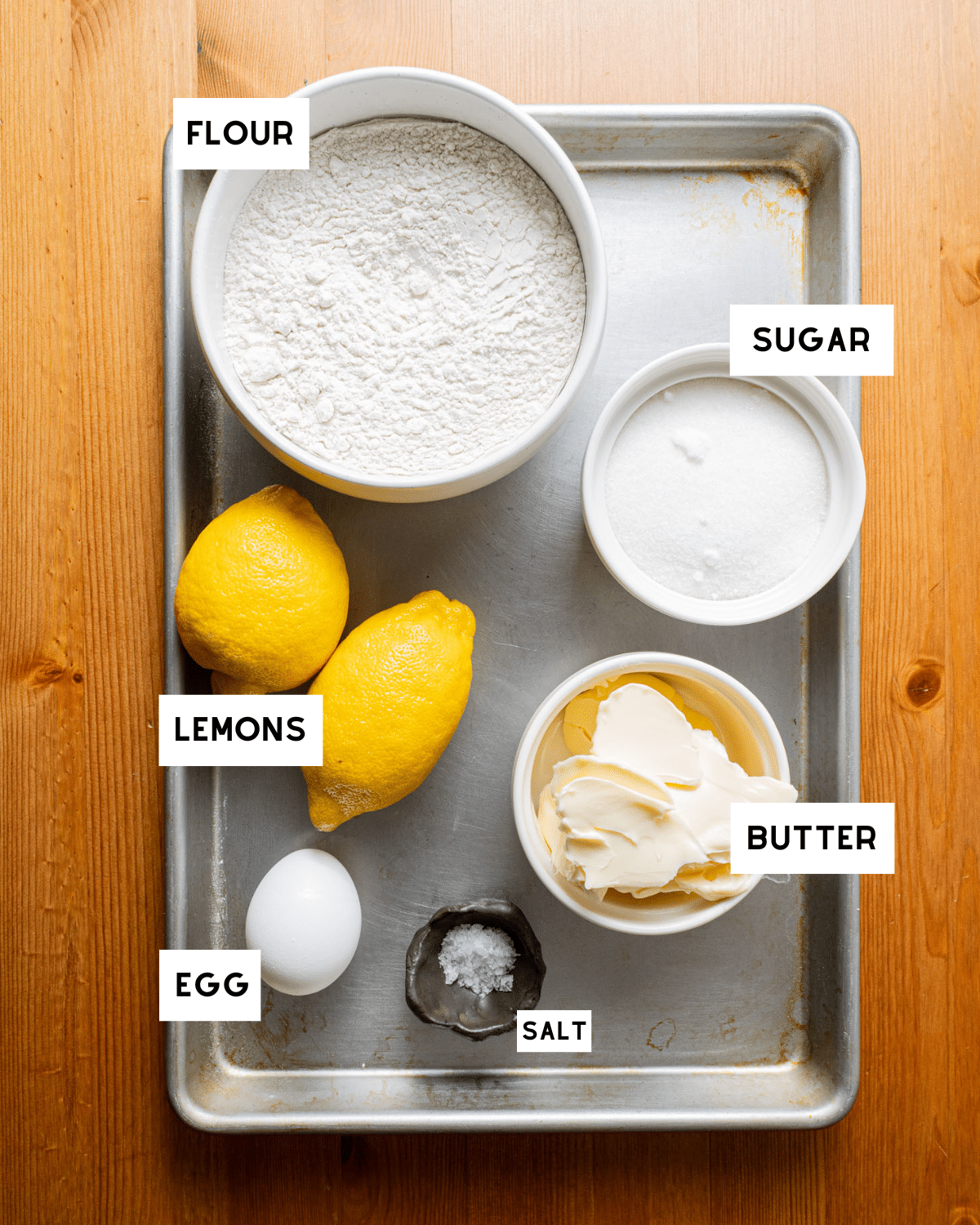 Baking ingredients on a tray: bowl of flour, sugar, two lemons, butter, an egg, and a pinch of salt. Labels identify each ingredient.