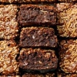 Rich chocolate brownies topped with golden toasted coconut, arranged in a grid. Three pieces are stacked, showing a moist, fudgy interior.