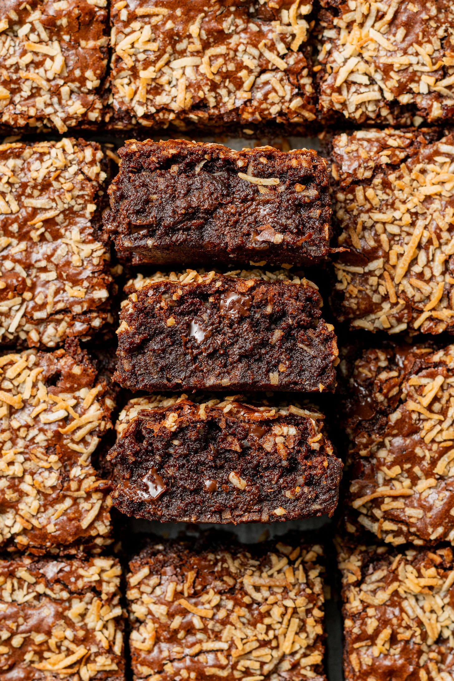 Rich chocolate brownies topped with golden toasted coconut, arranged in a grid. Three pieces are stacked, showing a moist, fudgy interior.