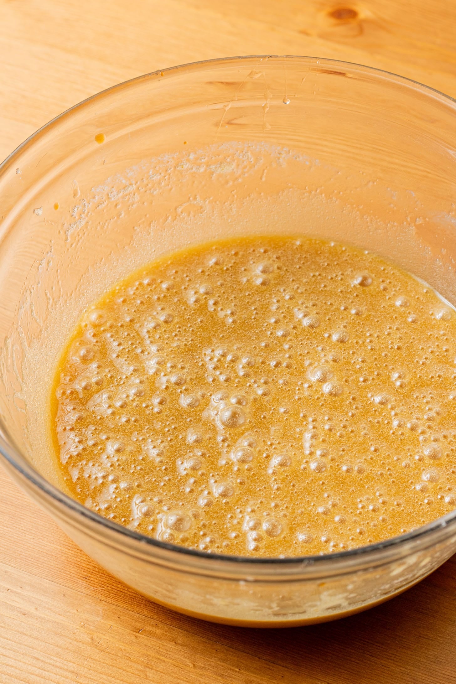 A glass bowl filled with frothy, light brown batter sits on a wooden surface. The mixture appears smooth with bubbles, suggesting active yeast.