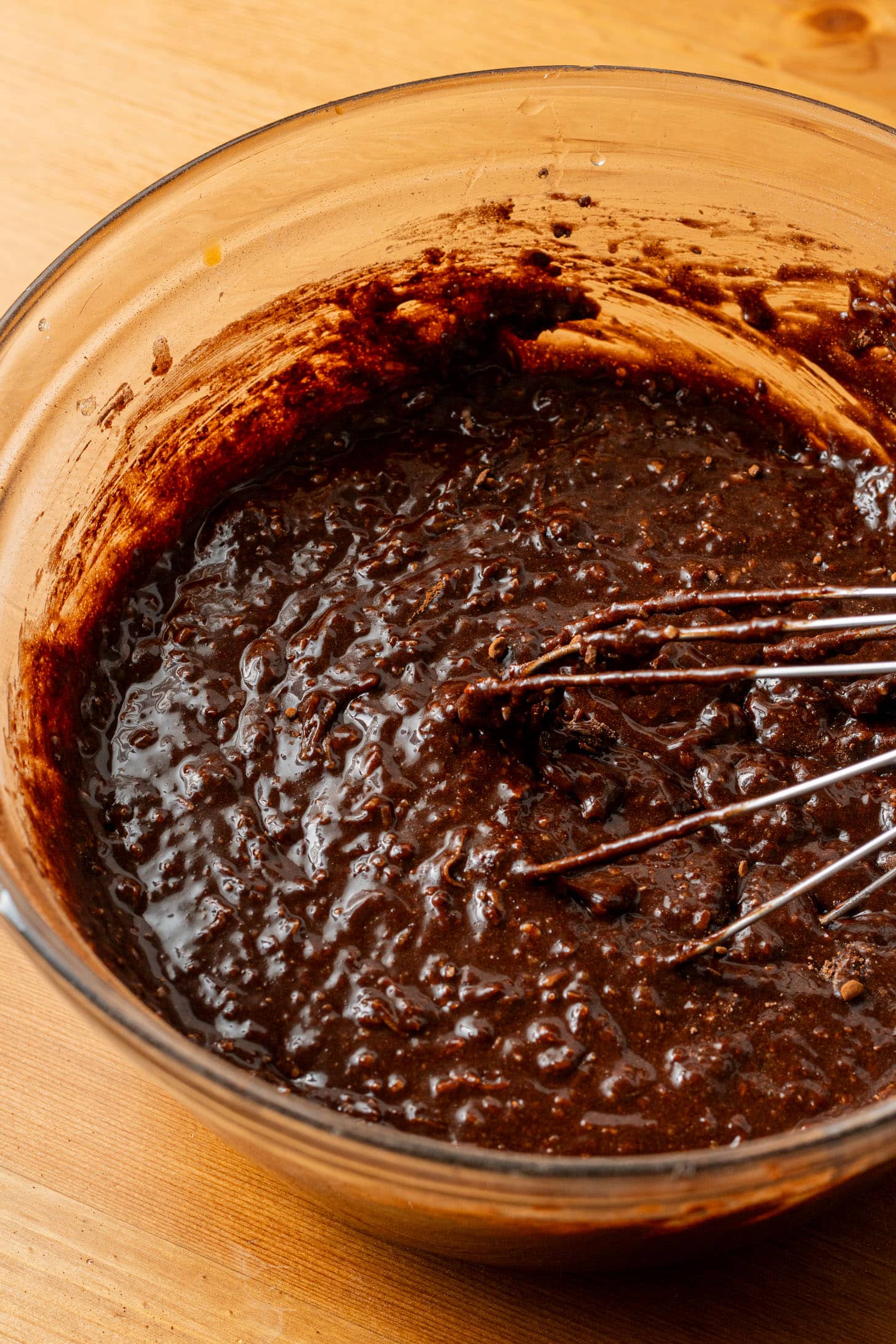 A glass bowl filled with thick, rich chocolate cake batter sits on a wooden table. A metal whisk rests inside, partially coated with batter.