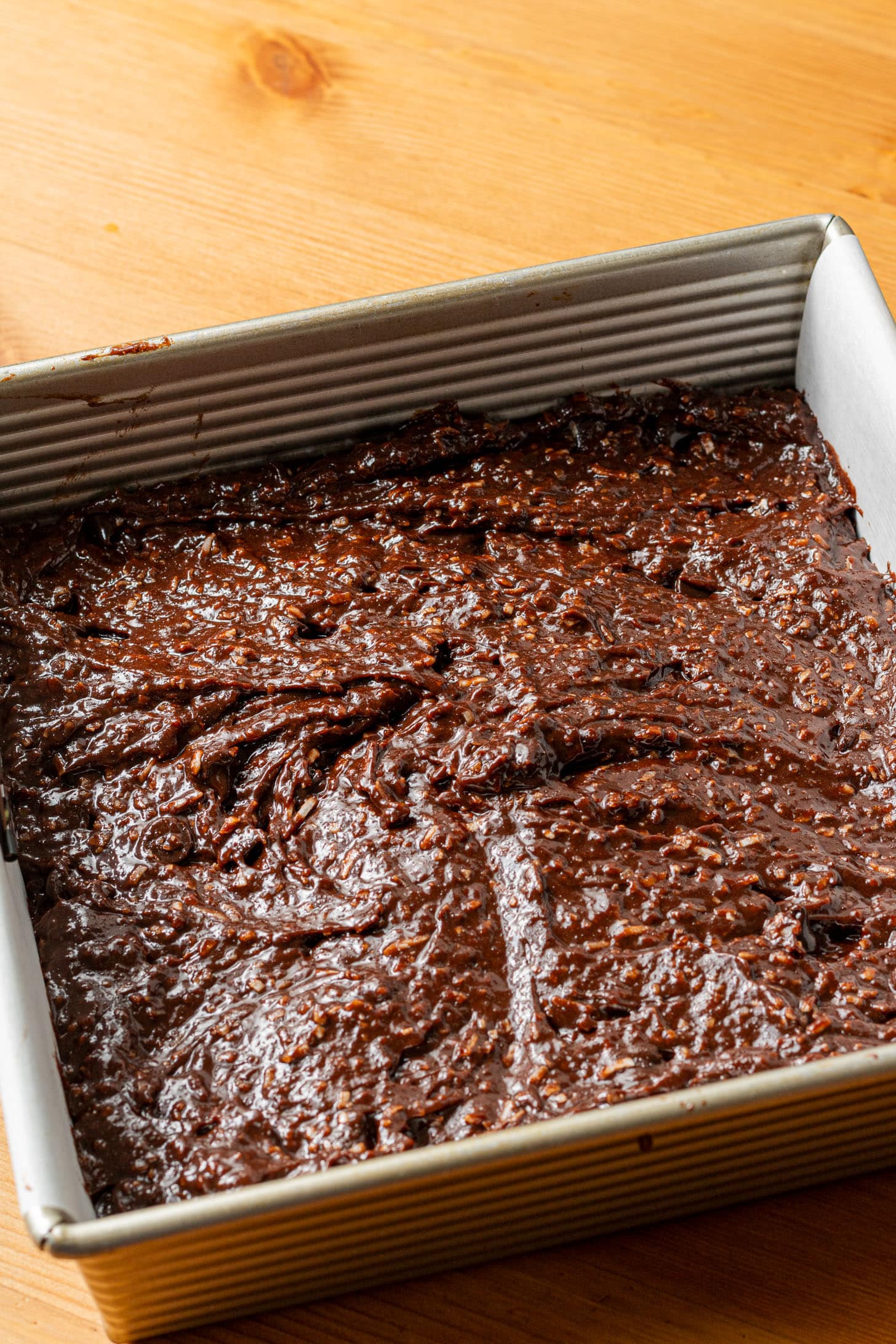 A square baking pan filled with rich, glossy chocolate brownie batter sits on a wooden table, exuding a deliciously indulgent and homemade feel.