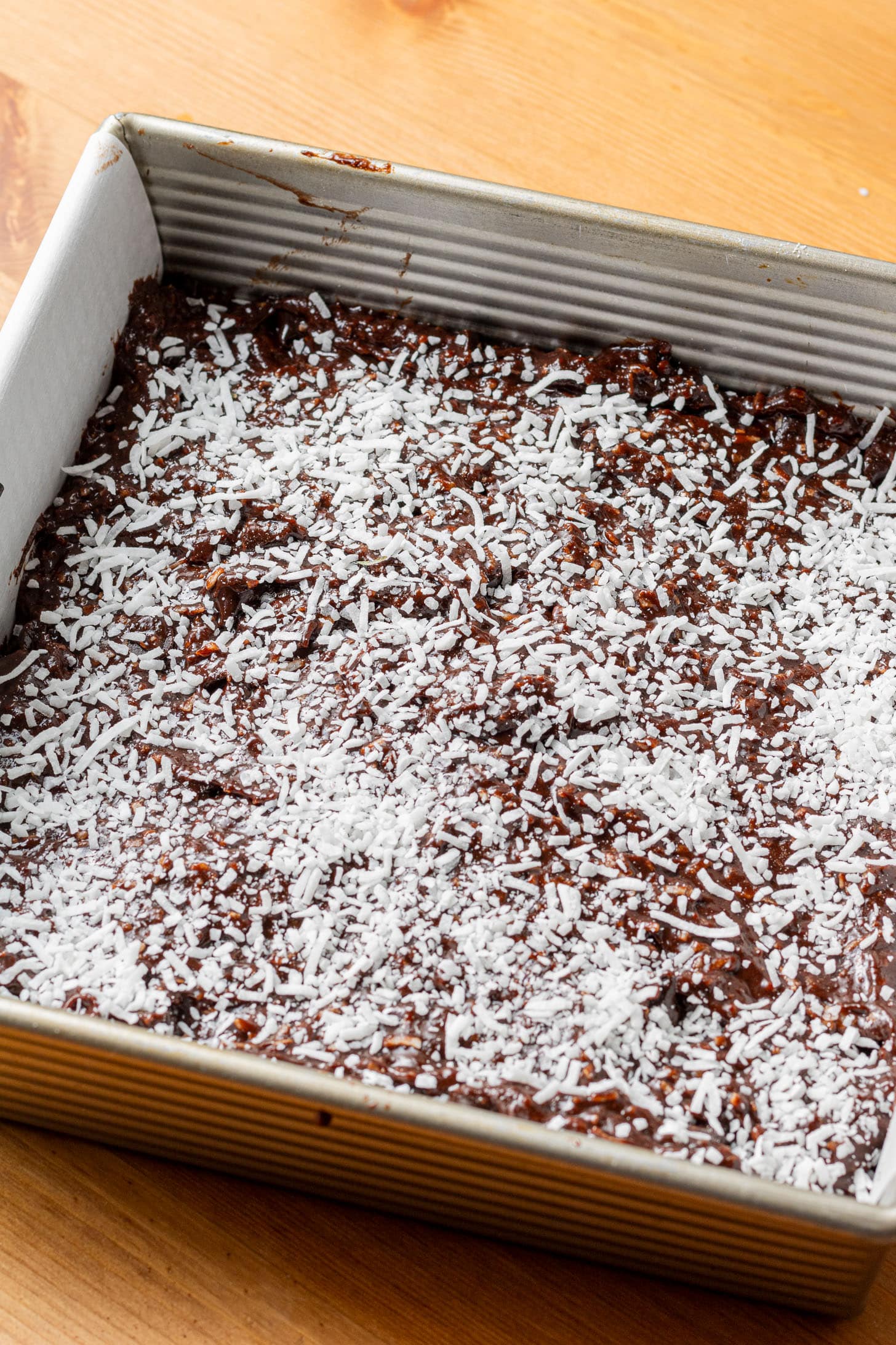A square baking pan filled with rich, moist chocolate cake topped with a generous layer of shredded coconut, placed on a wooden table.