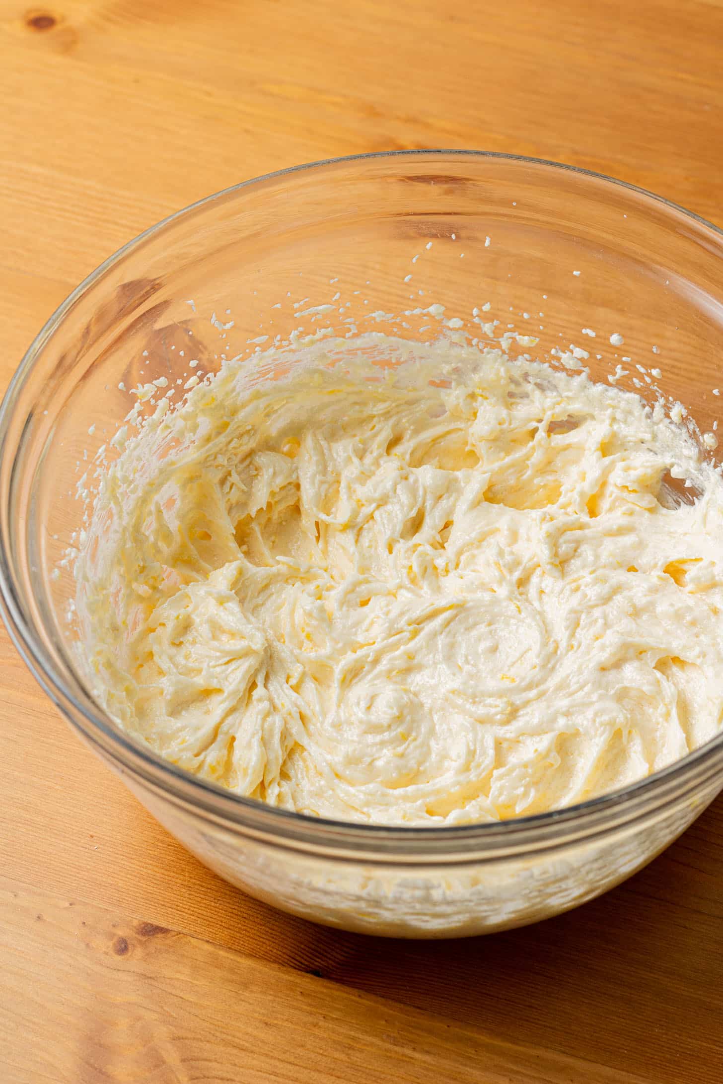 A glass bowl filled with creamy, light-yellow whipped butter on a wooden table, evoking a sense of anticipation and warmth in baking preparations.