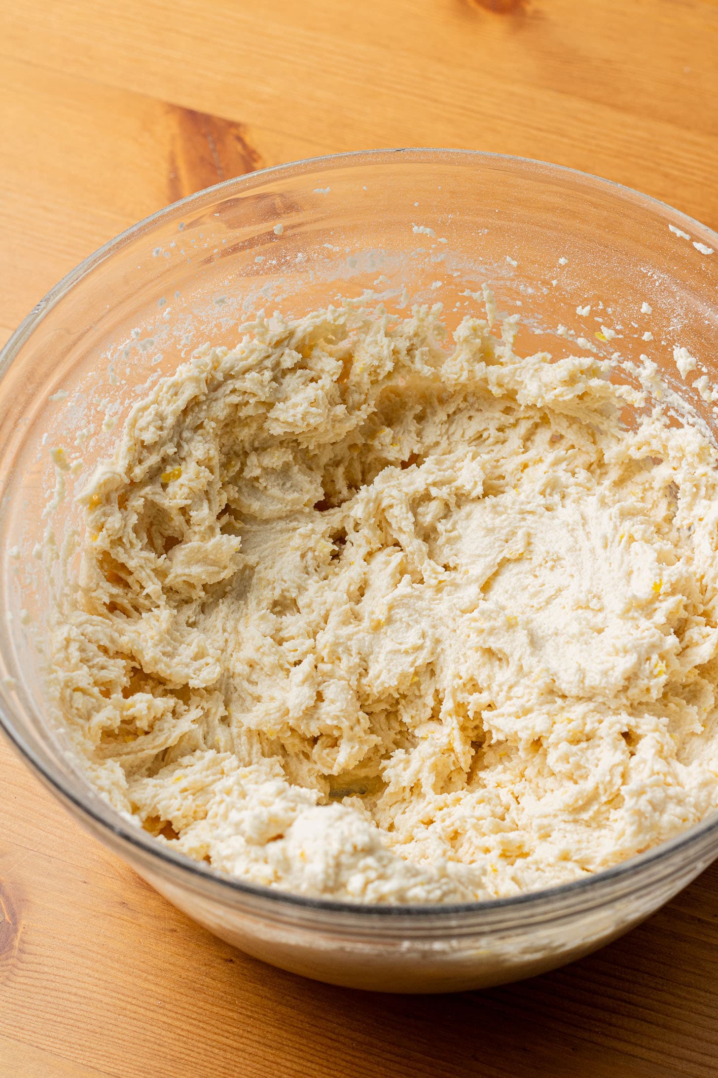 A glass bowl on a wooden surface holds creamy, mixed dough, suggesting a baking activity. The dough's texture is soft and slightly lumpy.