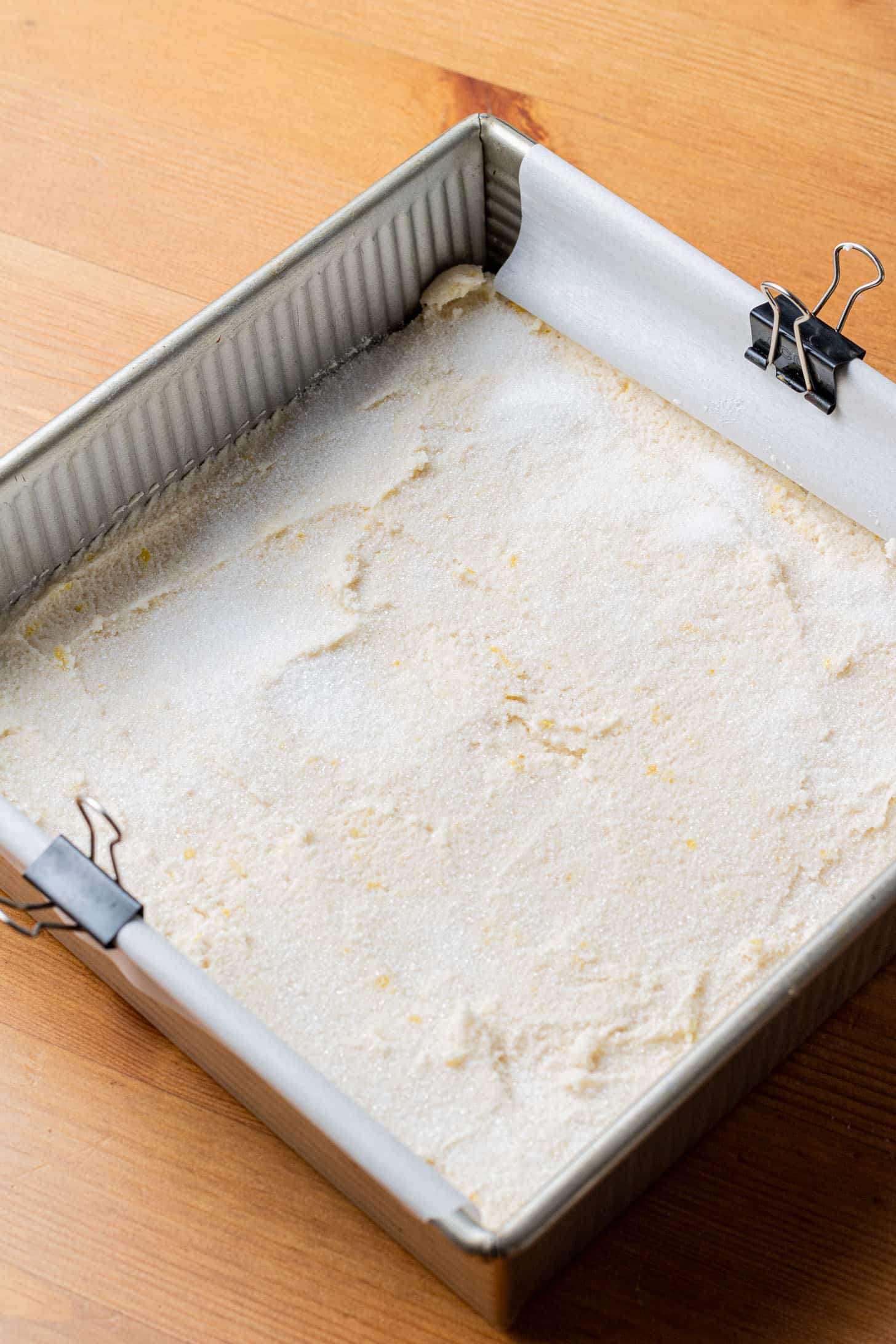 A metal baking pan lined with parchment paper, held by clips, contains an uncooked batter evenly spread. It sits on a wooden surface, ready for baking.