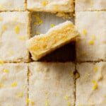 Close-up of lemon sugar cookie bars cut into squares, topped with lemon zest. One bar is slightly lifted, revealing a soft, crumbly texture.