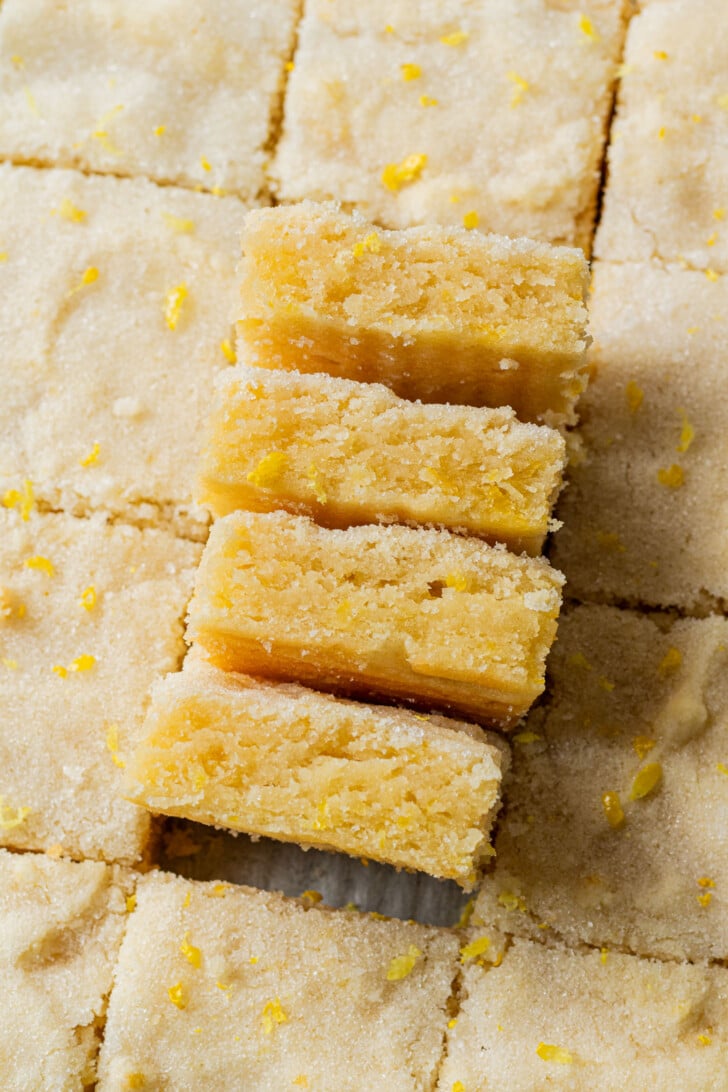 Close-up of lemon bars arranged in a grid, with a stack of four bars slightly offset. Each bar is flecked with lemon zest, conveying a fresh, homemade feel.