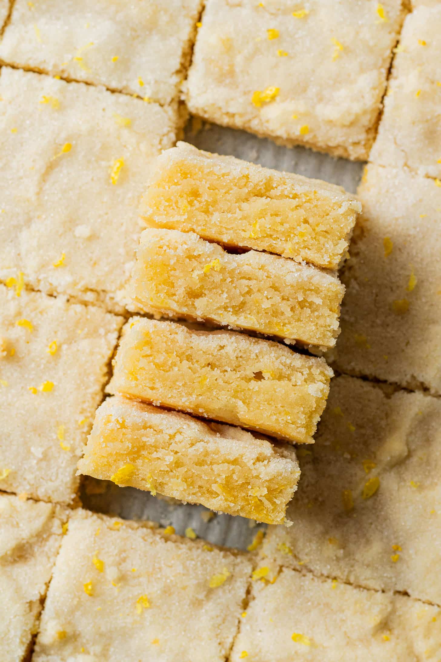 Close-up of lemon bars arranged in a grid, with a stack of four bars slightly offset. Each bar is flecked with lemon zest, conveying a fresh, homemade feel.