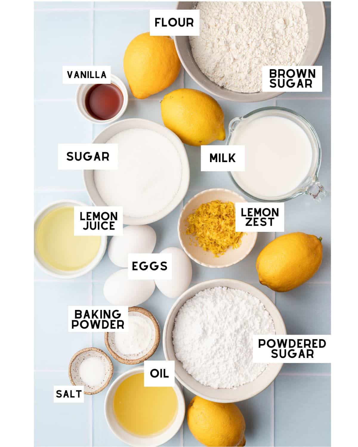 Assorted baking ingredients on a light blue surface labeled: flour, brown sugar, milk, vanilla, eggs, oil, lemon juice, lemon zest, powdered sugar, salt, baking powder. Lemons and sugar are also visible.
