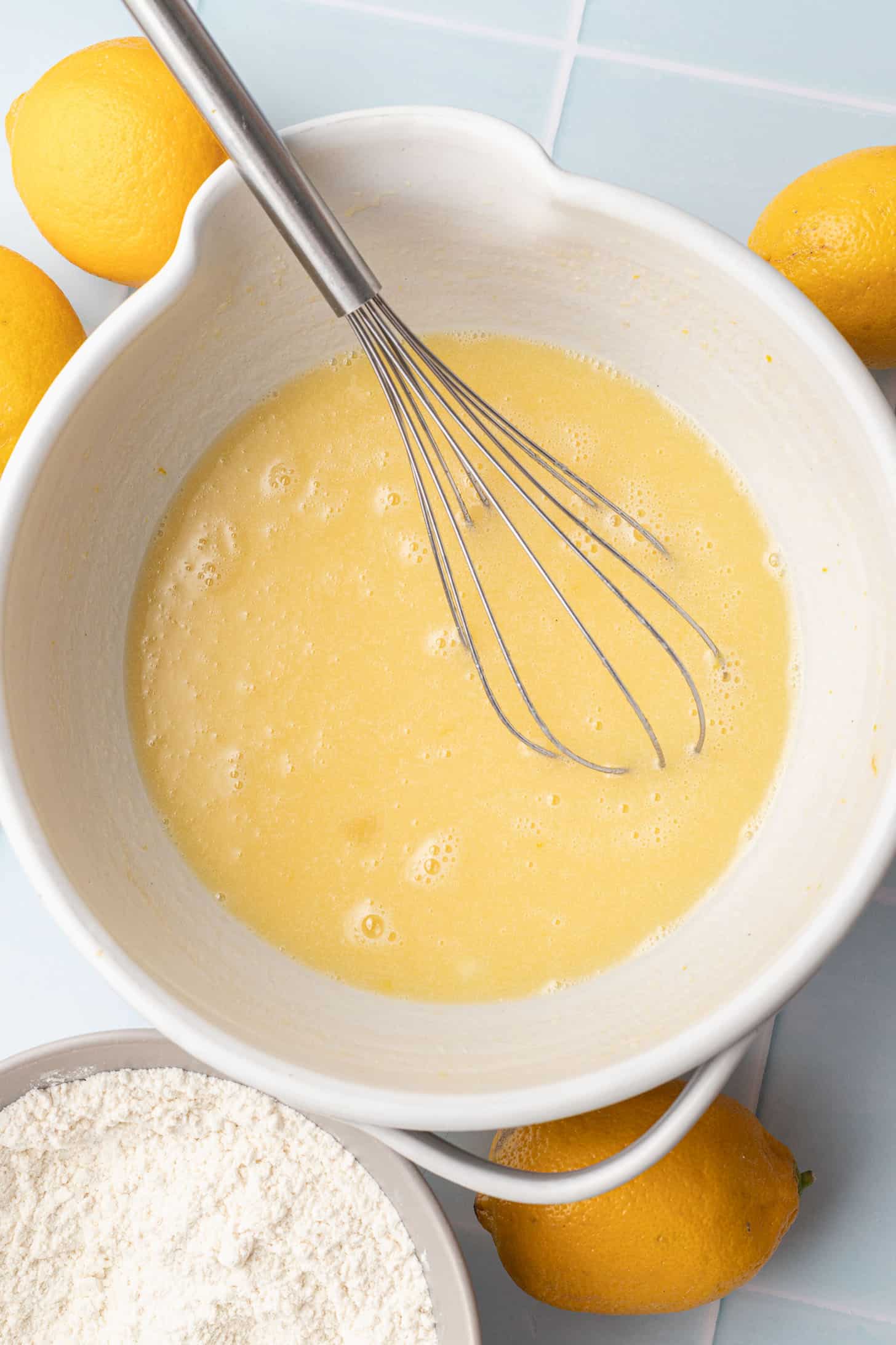 A white mixing bowl with yellow batter and a whisk inside, surrounded by fresh lemons and a bowl of flour, set on a light blue tiled surface.