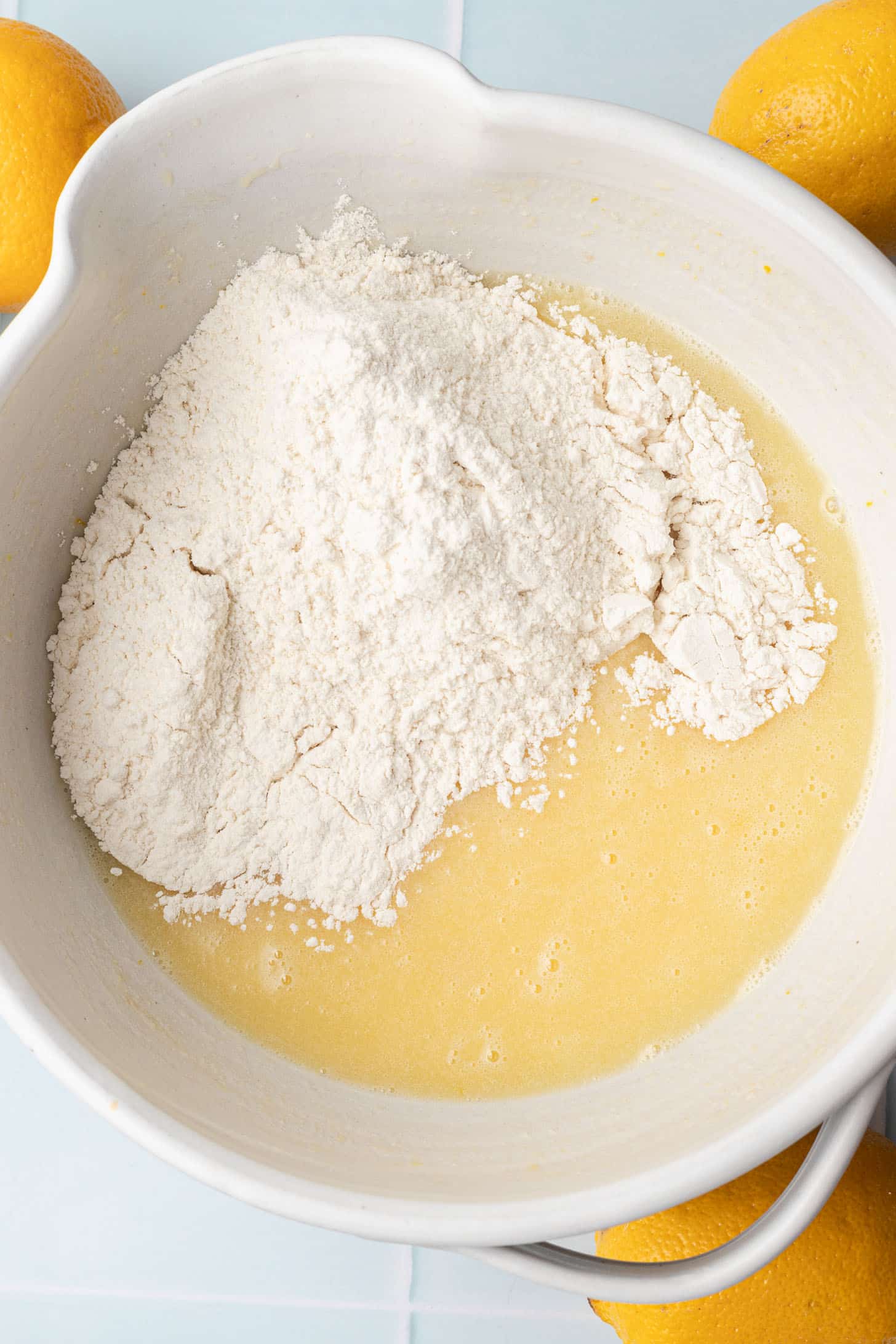 A white mixing bowl contains flour and yellow batter, ready to be mixed. Bright lemons surround the bowl, suggesting a lemon-flavored recipe.