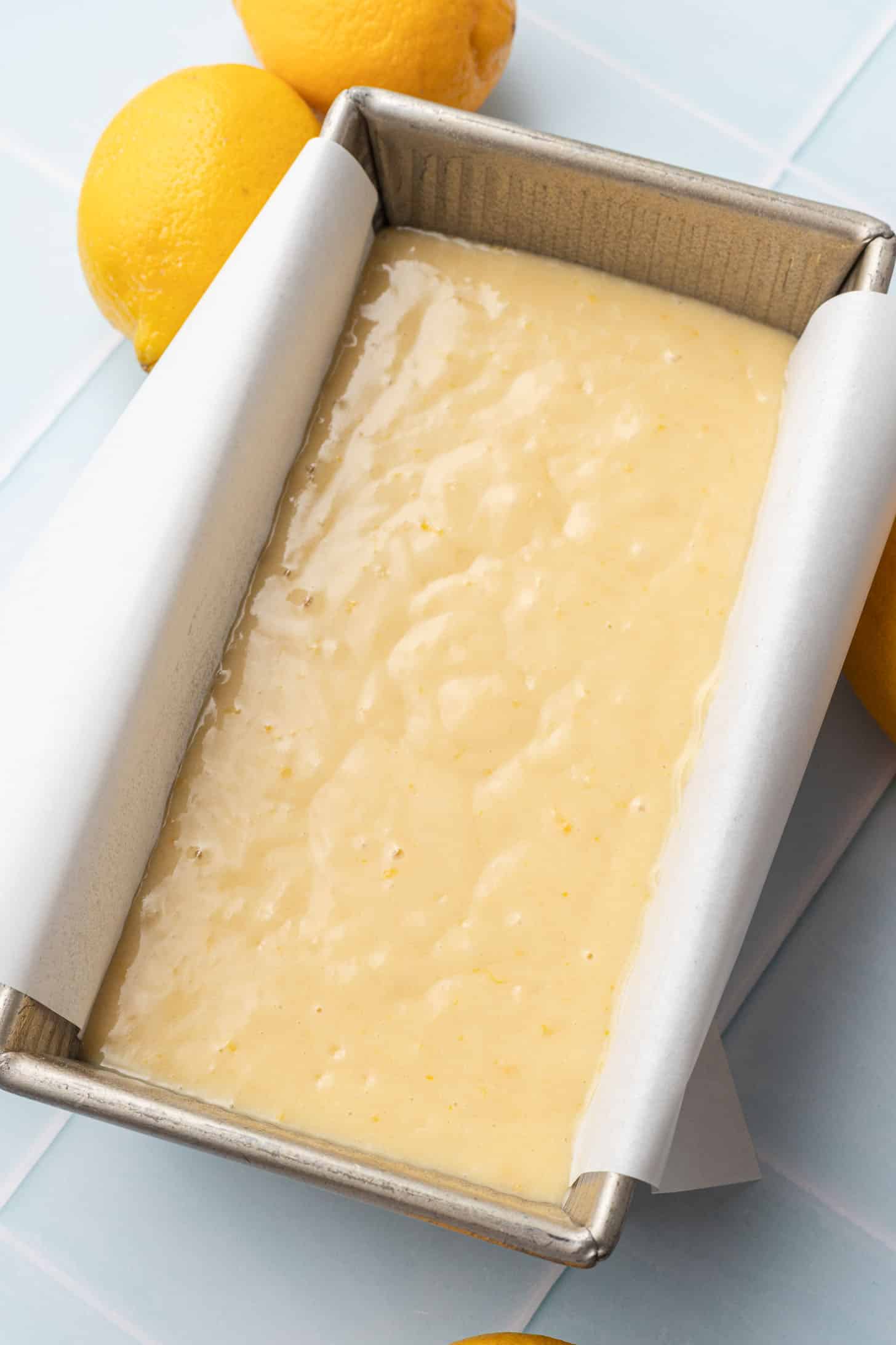 Lemon cake batter in a parchment-lined rectangular pan, surrounded by fresh lemons on a light blue surface, suggesting a fresh, zesty flavor.