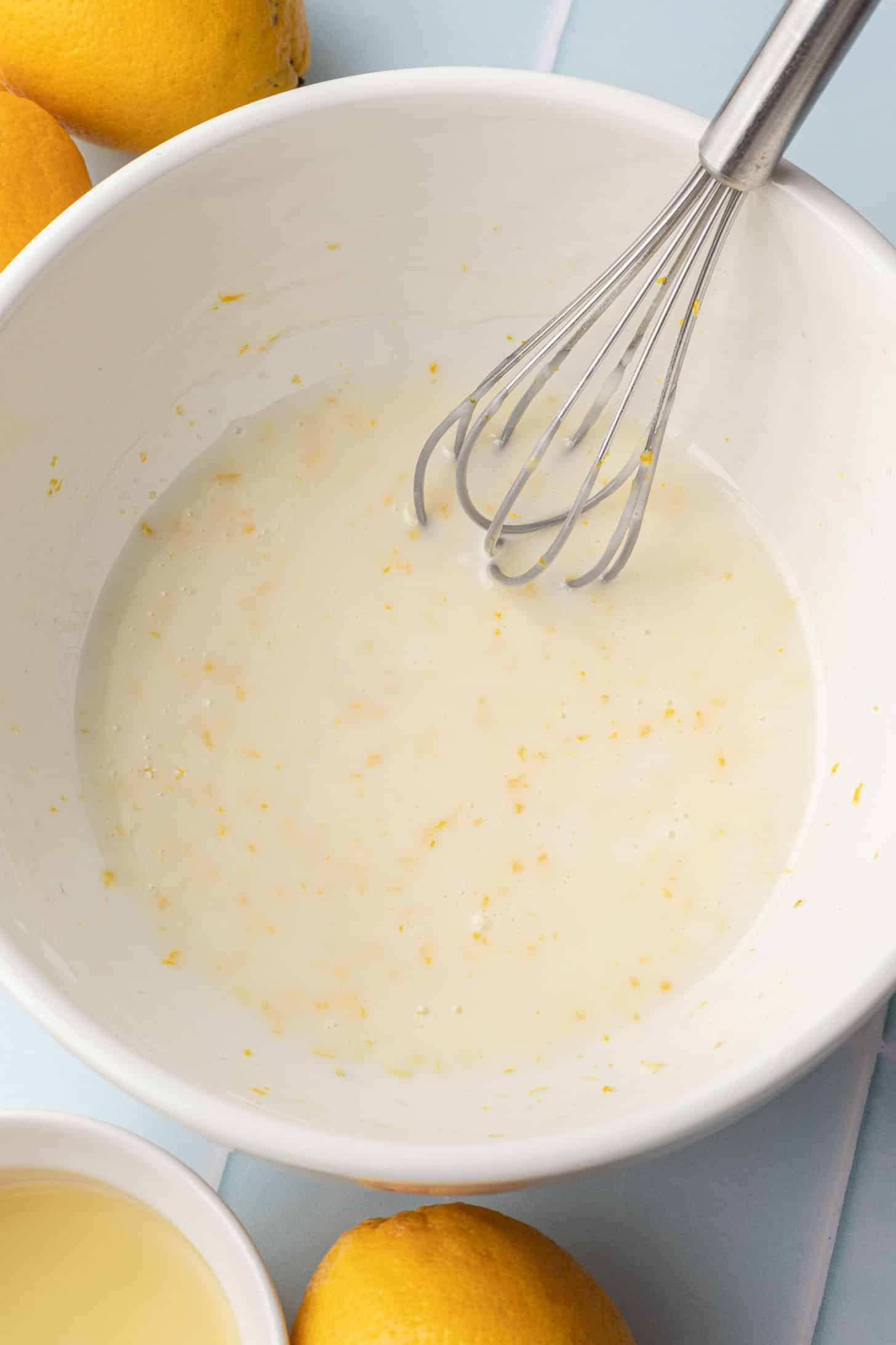 A whisk in a white bowl with lemon zest-speckled glaze mixture, surrounded by fresh lemons on a light blue surface, conveying freshness and zest.