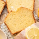 Close-up of sliced lemon cake on a cooling rack, with a glaze and lemon slices on top. Fresh lemons and a bowl are nearby, creating a warm, inviting feel.