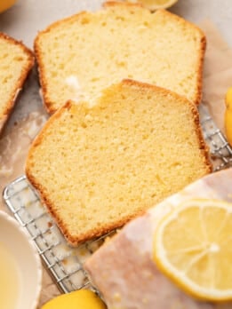 Close-up of sliced lemon cake on a cooling rack, with a glaze and lemon slices on top. Fresh lemons and a bowl are nearby, creating a warm, inviting feel.