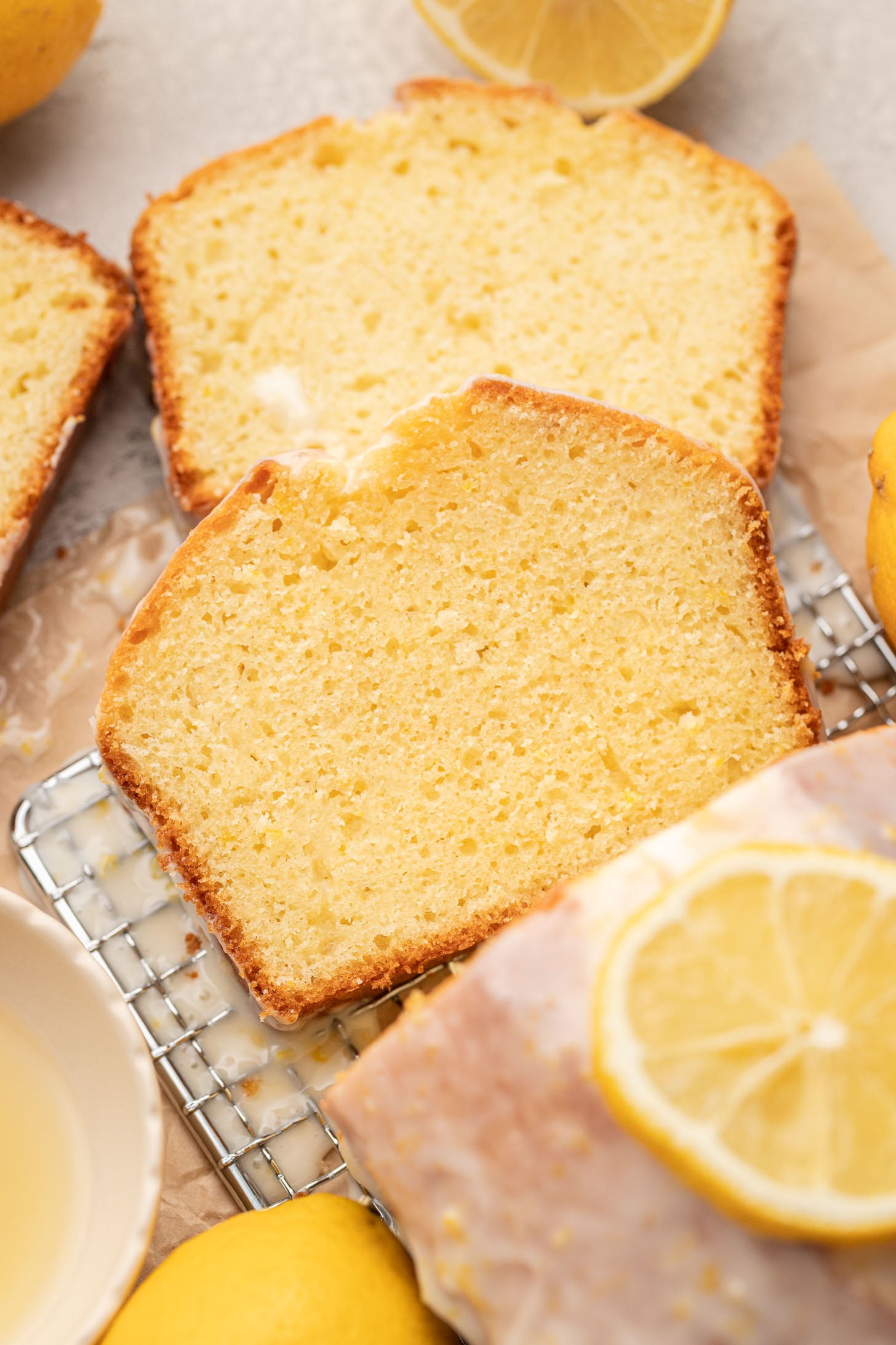 Close-up of sliced lemon cake on a cooling rack, with a glaze and lemon slices on top. Fresh lemons and a bowl are nearby, creating a warm, inviting feel.