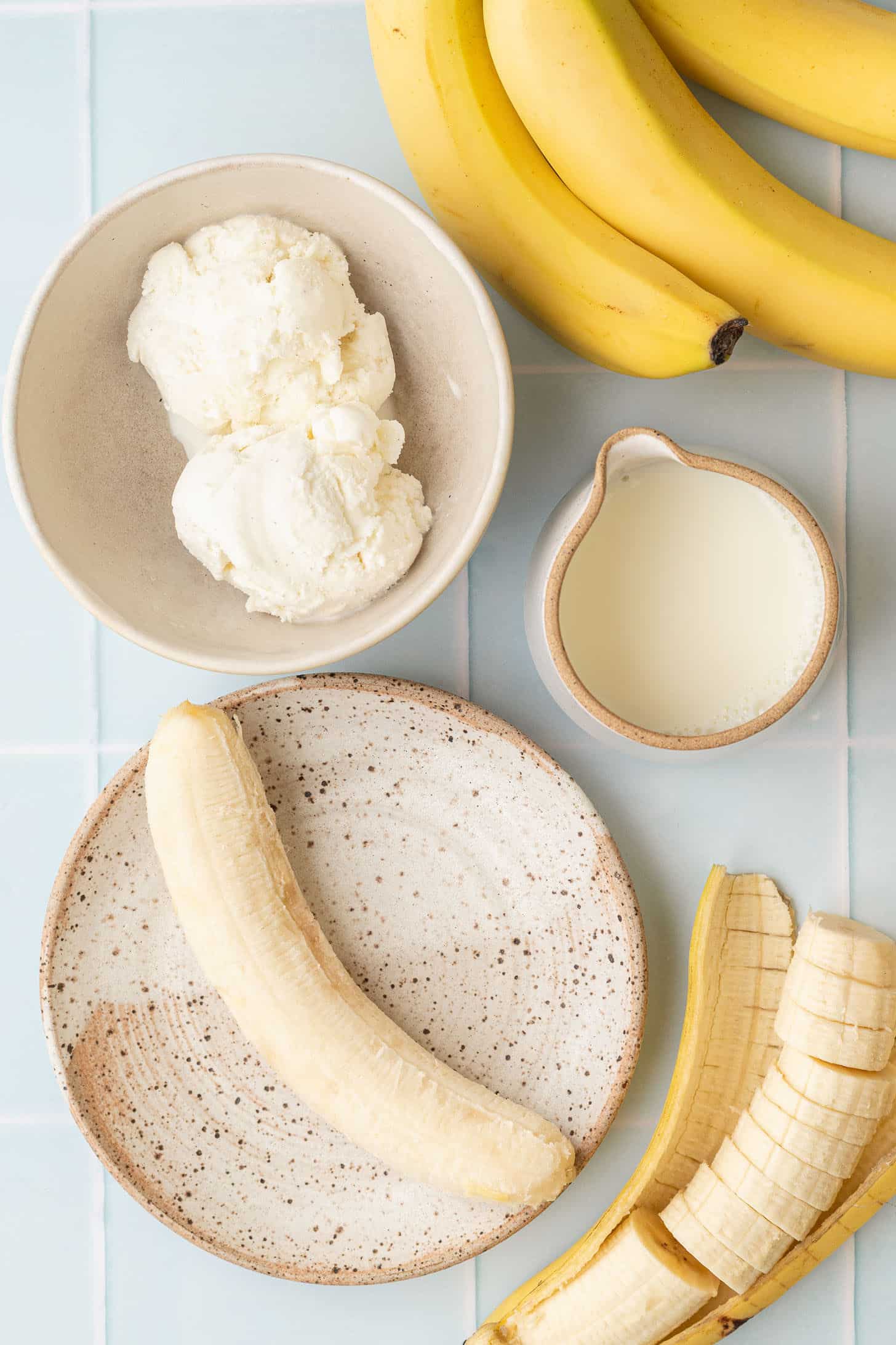 Flat lay of banana milkshake ingredients including scoops of vanilla ice cream, fresh bananas, a sliced banana, and a small pitcher of milk on a light blue tile surface
