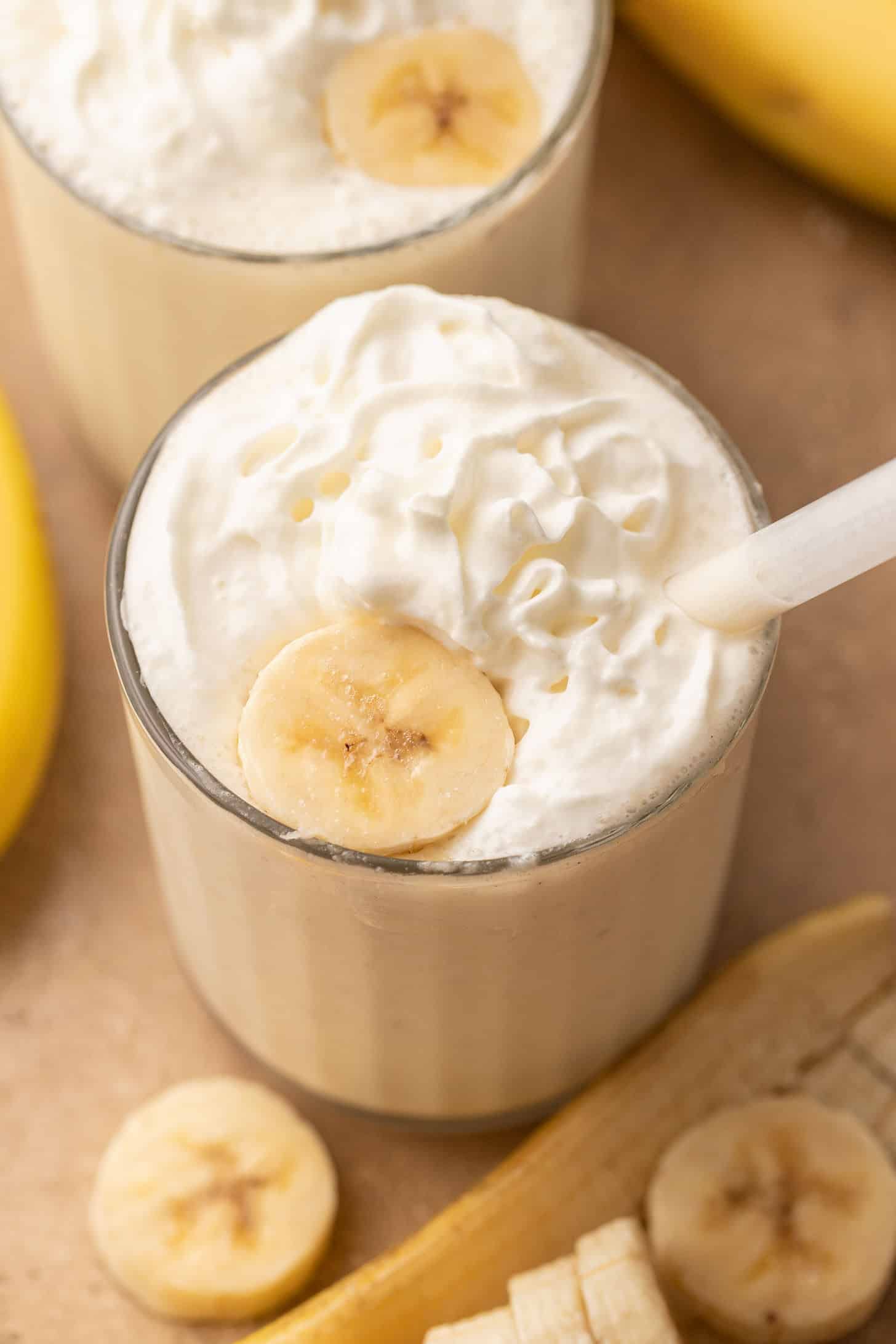 Two banana milkshakes in clear glasses topped with whipped cream and a banana slice, with a white straw, surrounded by sliced bananas and whole bananas on a wooden surface