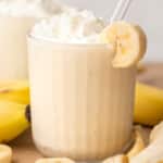 A banana milkshake in a clear glass topped with whipped cream and a banana slice on the rim with a white straw, surrounded by fresh banana slices and whole bananas on a wooden surface