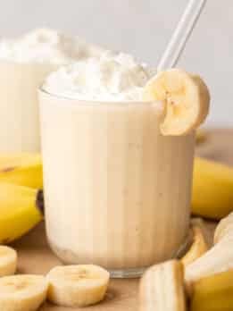 A banana milkshake in a clear glass topped with whipped cream and a banana slice on the rim with a white straw, surrounded by fresh banana slices and whole bananas on a wooden surface