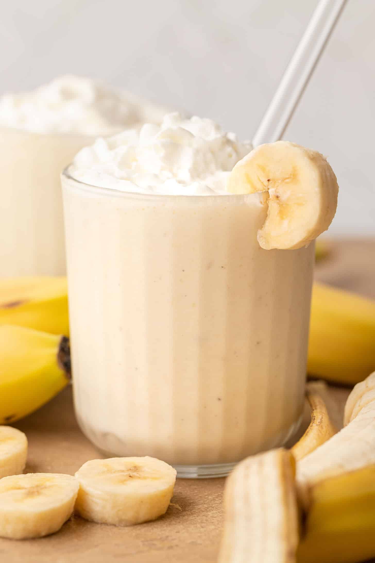 A banana milkshake in a clear glass topped with whipped cream and a banana slice on the rim with a white straw, surrounded by fresh banana slices and whole bananas on a wooden surface