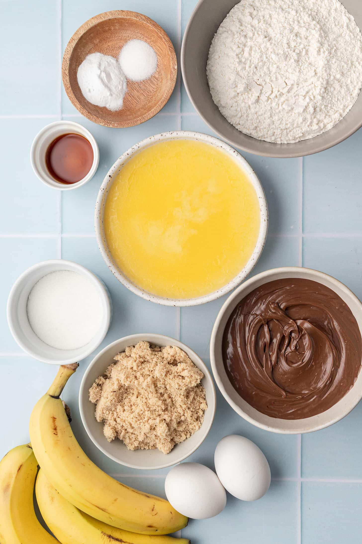 Overhead view of banana nutella muffin ingredients arranged on a light blue tile surface, including flour, melted butter, Nutella, brown sugar, white sugar, eggs, bananas, vanilla extract, and leavening agents