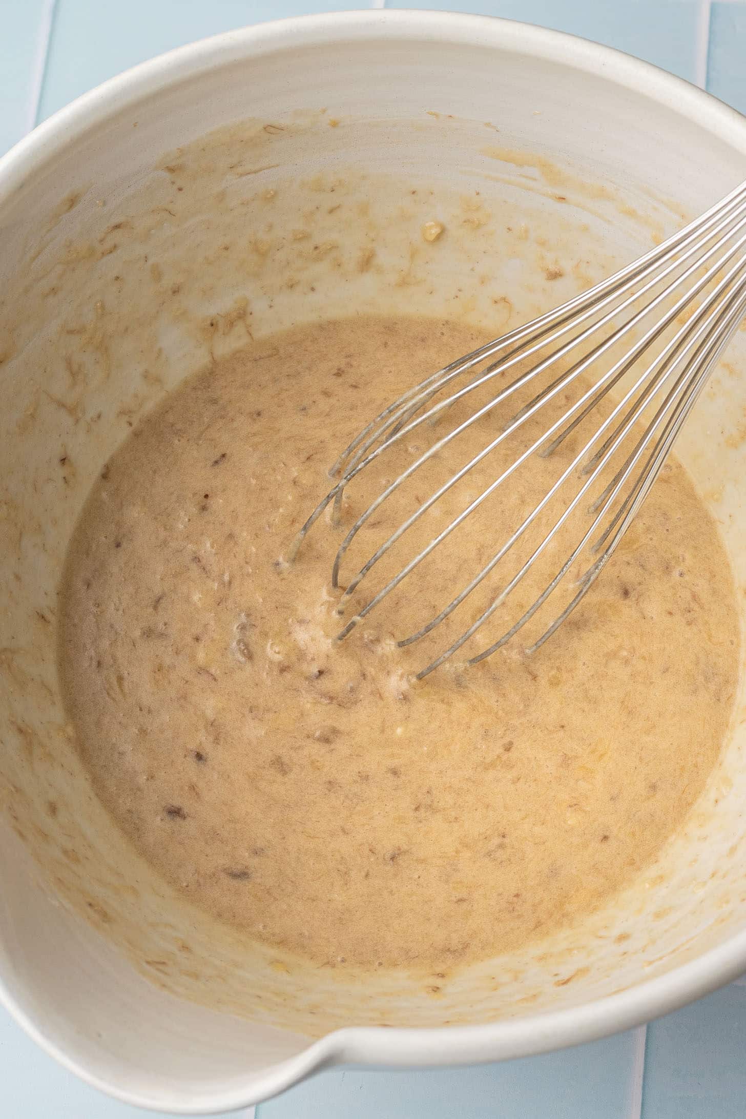 Overhead view of wet banana muffin batter being whisked in a large white ceramic bowl, showing a smooth, pale golden mixture with a stainless steel whisk