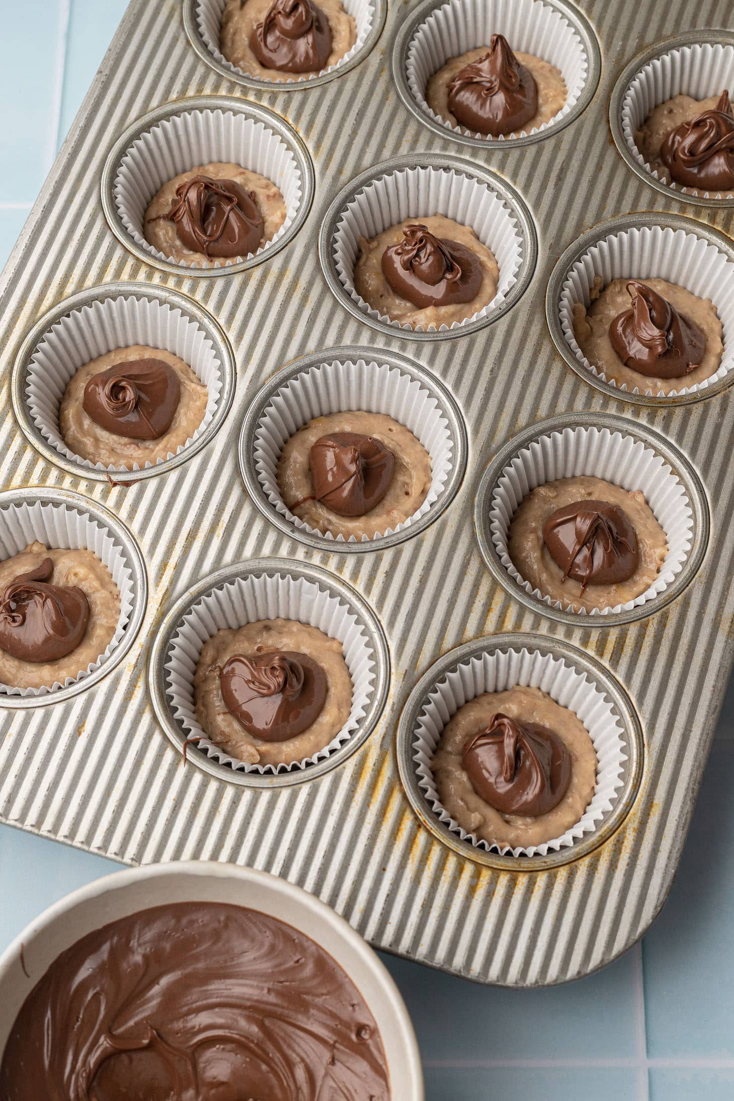 Overhead view of a silver muffin tin lined with white paper liners, each filled with banana batter and topped with a dollop of Nutella before swirling