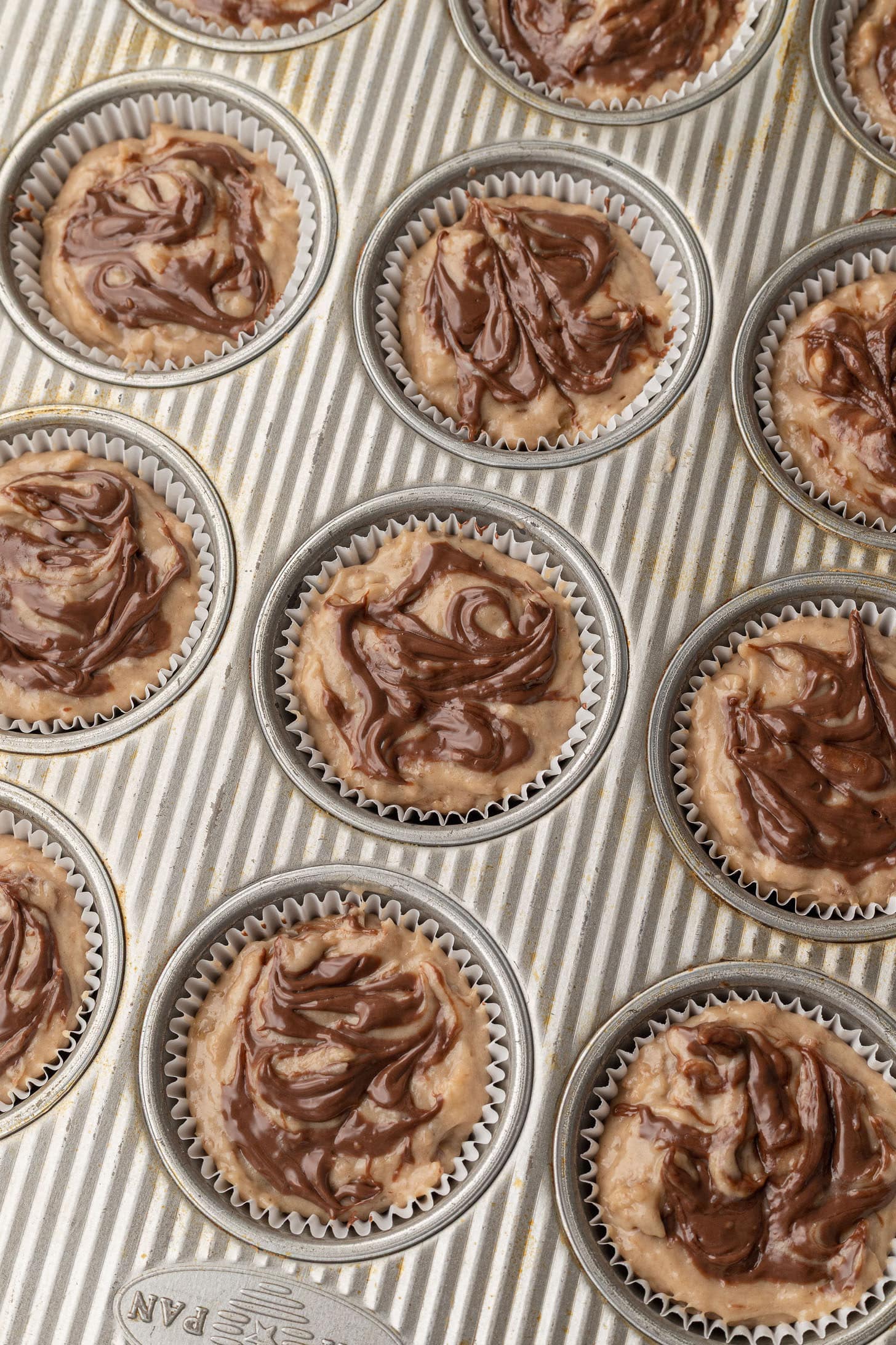 Close-up overhead view of muffin tin filled with banana batter swirled with Nutella in a marbled chocolate pattern, ready to go into the oven