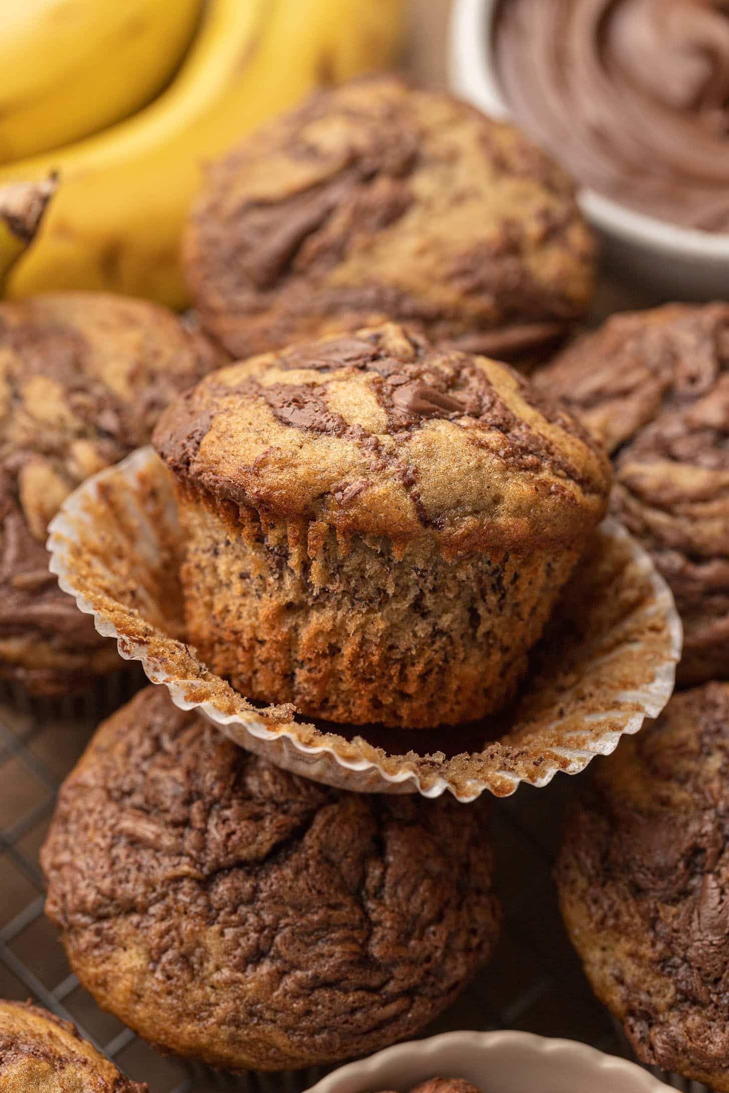 Close-up of two banana Nutella muffins stacked in a white paper liner, showing a golden brown domed top with chocolate swirls, surrounded by more muffins and bananas