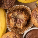 Close-up of a banana Nutella muffin with a bite taken out, revealing a warm gooey Nutella center, surrounded by more muffins, fresh bananas, and a small bowl of Nutella in the background