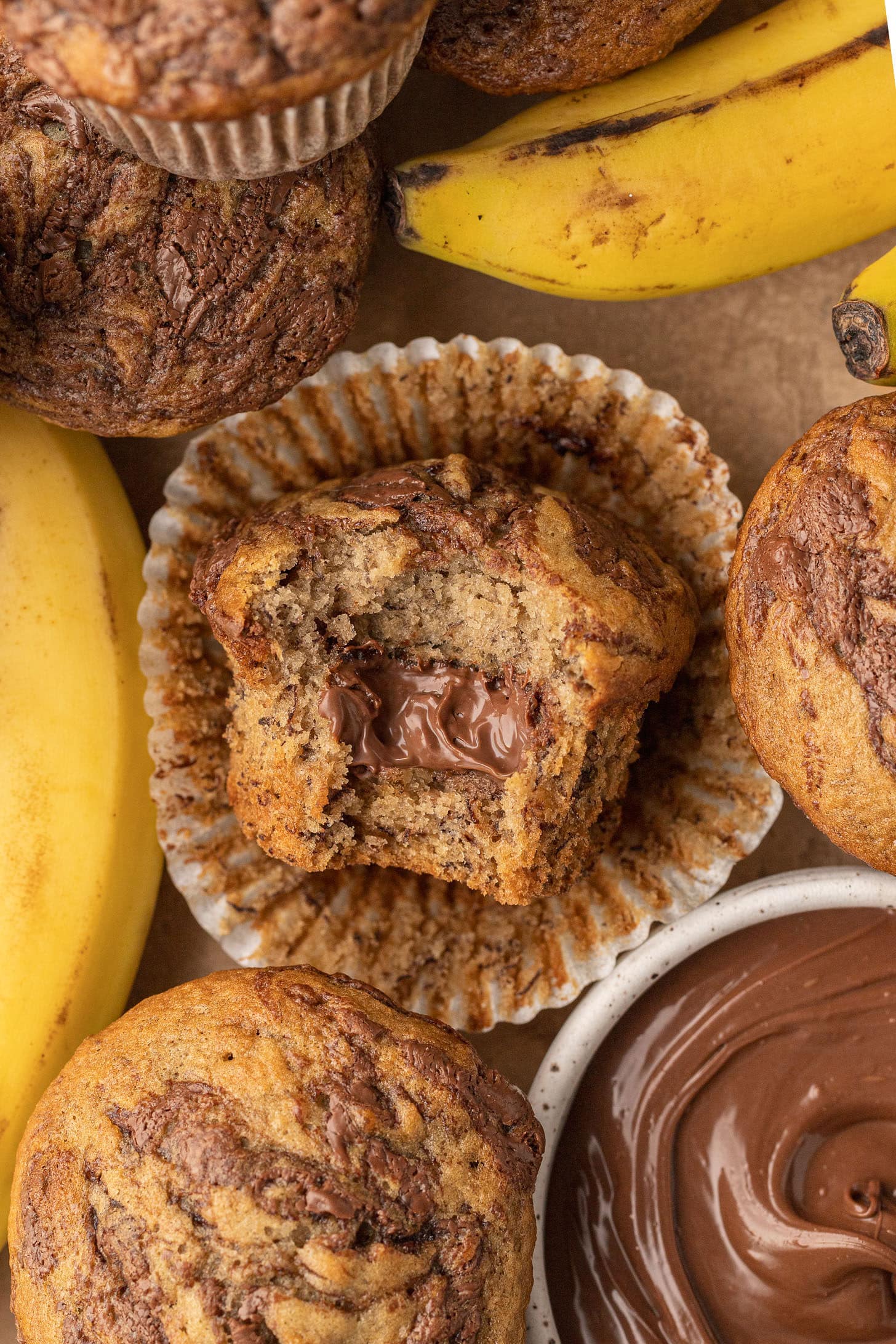 Close-up of a banana Nutella muffin with a bite taken out, revealing a warm gooey Nutella center, surrounded by more muffins, fresh bananas, and a small bowl of Nutella in the background