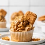 A cupcake topped with a generous swirl of Biscoff buttercream, drizzled with cookie butter, sprinkled with Biscoff cookie crumbs, and garnished with a whole Lotus Biscoff cookie, served on a white plate